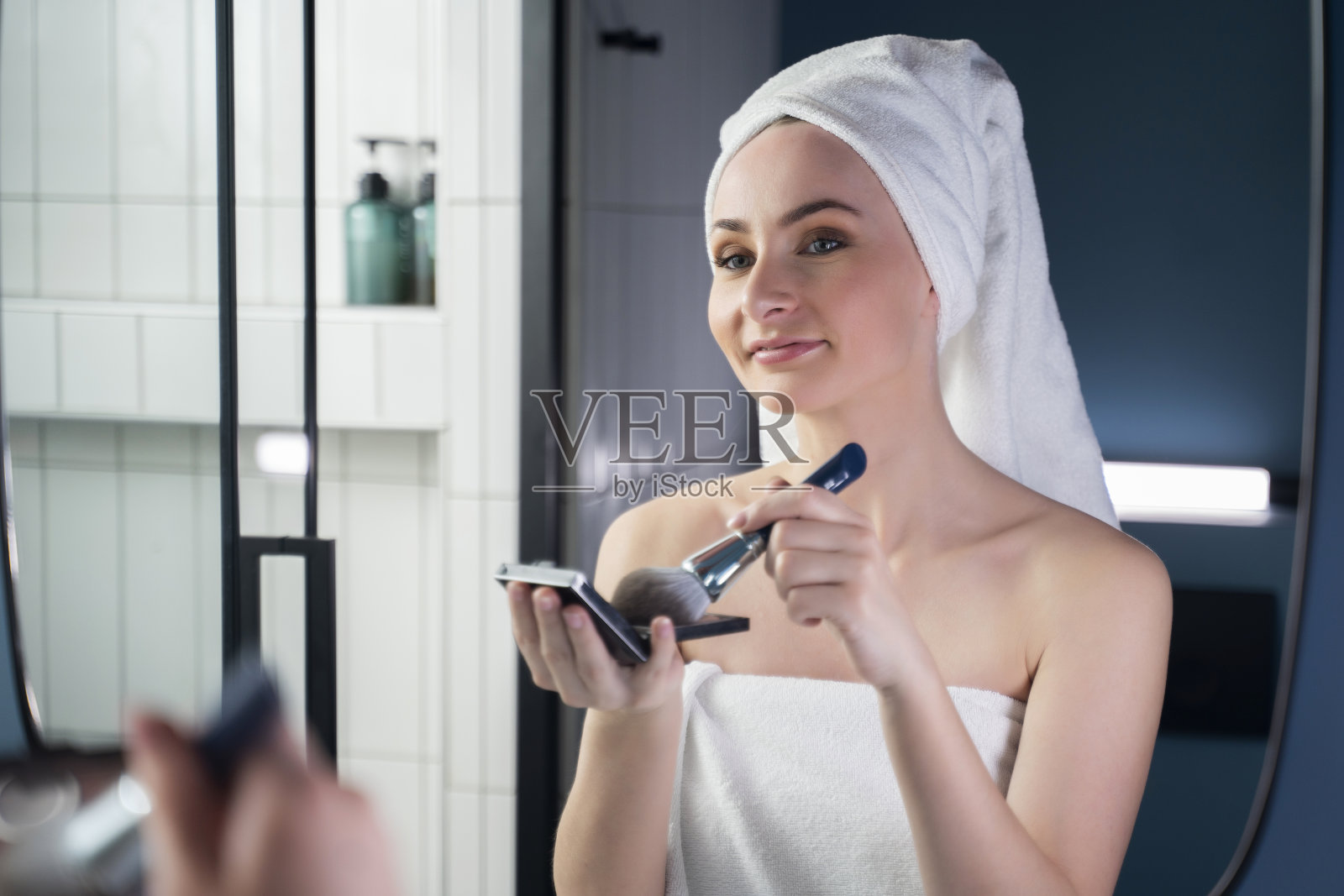 一名年轻女子在浴室里用毛巾蒙头，拿着腮红刷，为晚妆涂粉底照片摄影图片