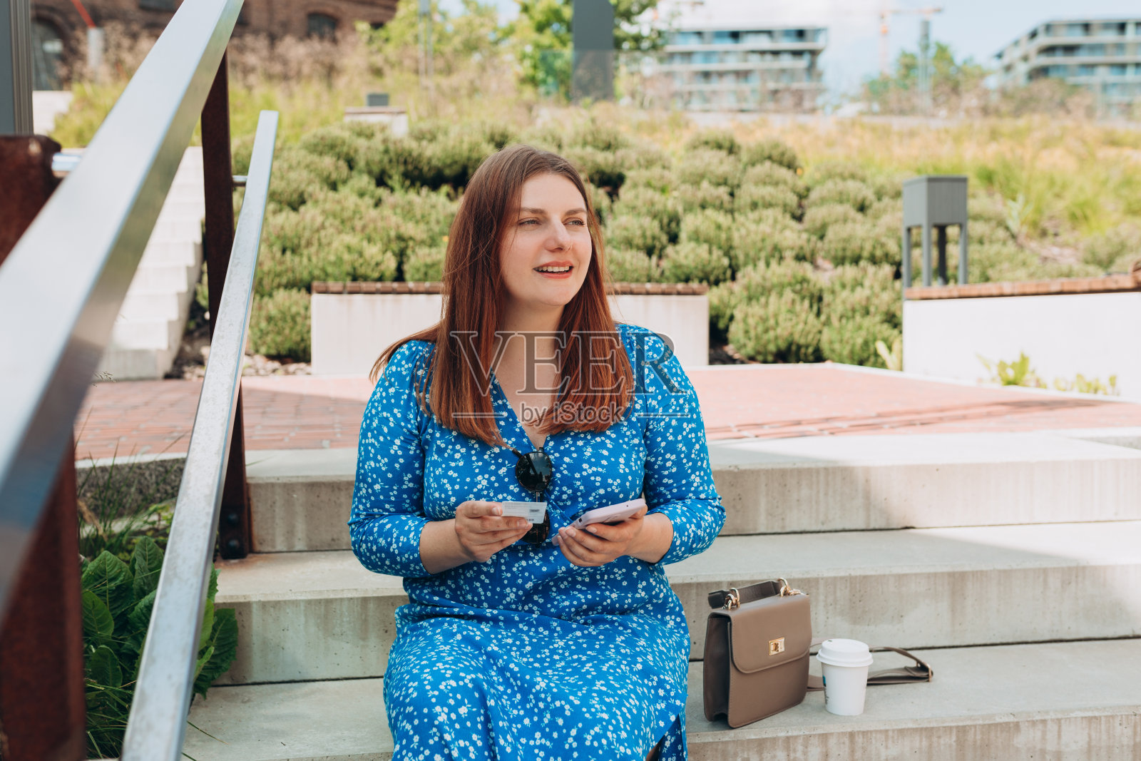 非常快乐的红发女人站在城市背景上，手里拿着车票和手机。女人手里拿着公交或地铁车票，女孩坐在城市街道的楼梯上。照片摄影图片