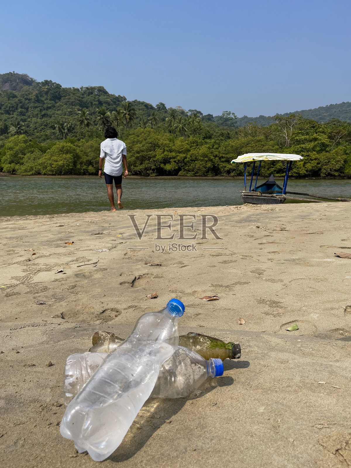 特写照片:退潮时被冲到沙滩上的三堆塑料水瓶，海岸上的海洋垃圾和污染，乱扔的沙子，肮脏的海滩，印度人站在渔船边的海边，有棕榈树的林地照片摄影图片