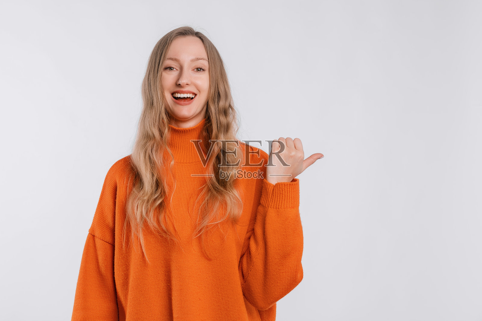 积极的年轻女子用手指向右，展示广告或横幅，愉快地微笑着，穿着一件柔软的橙色毛衣，站在中性的背景上。好主意，好提议照片摄影图片