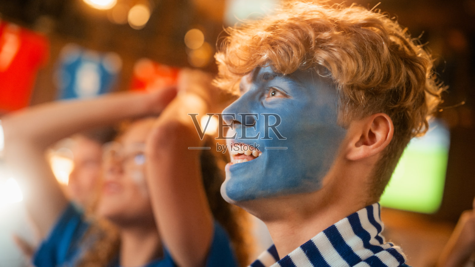 近距离拍摄一群脸涂成蓝白相间的球迷站在酒吧里为他们的球队加油。举手呐喊。朋友们在进球后庆祝胜利。照片摄影图片