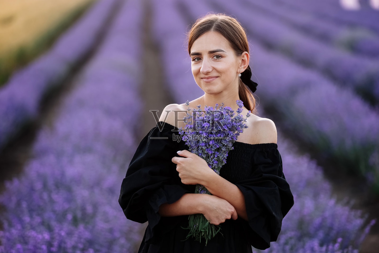 年轻浪漫的女人，健康的自然美丽的皮肤，拿着花束在薰衣草田看着相机，微笑。漂亮的女孩穿着黑色的裙子，头上戴着蝴蝶结。天然产品广告照片摄影图片
