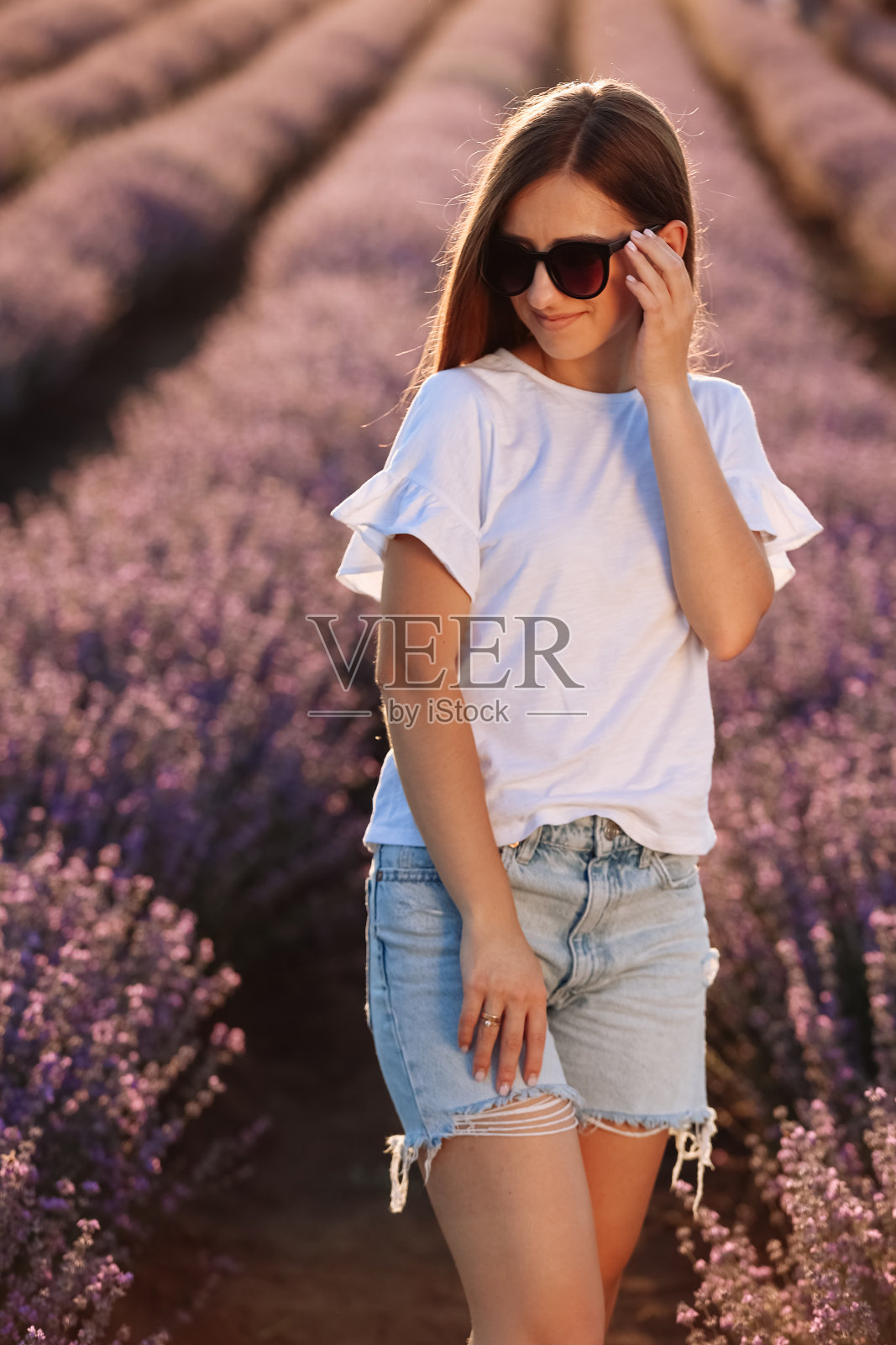 美丽的年轻女子走在夕阳下，看着薰衣草田。戴着墨镜、穿着便服的女孩站在紫色的薰衣草地里。享受花香的林间空地，夏日自然。照片摄影图片