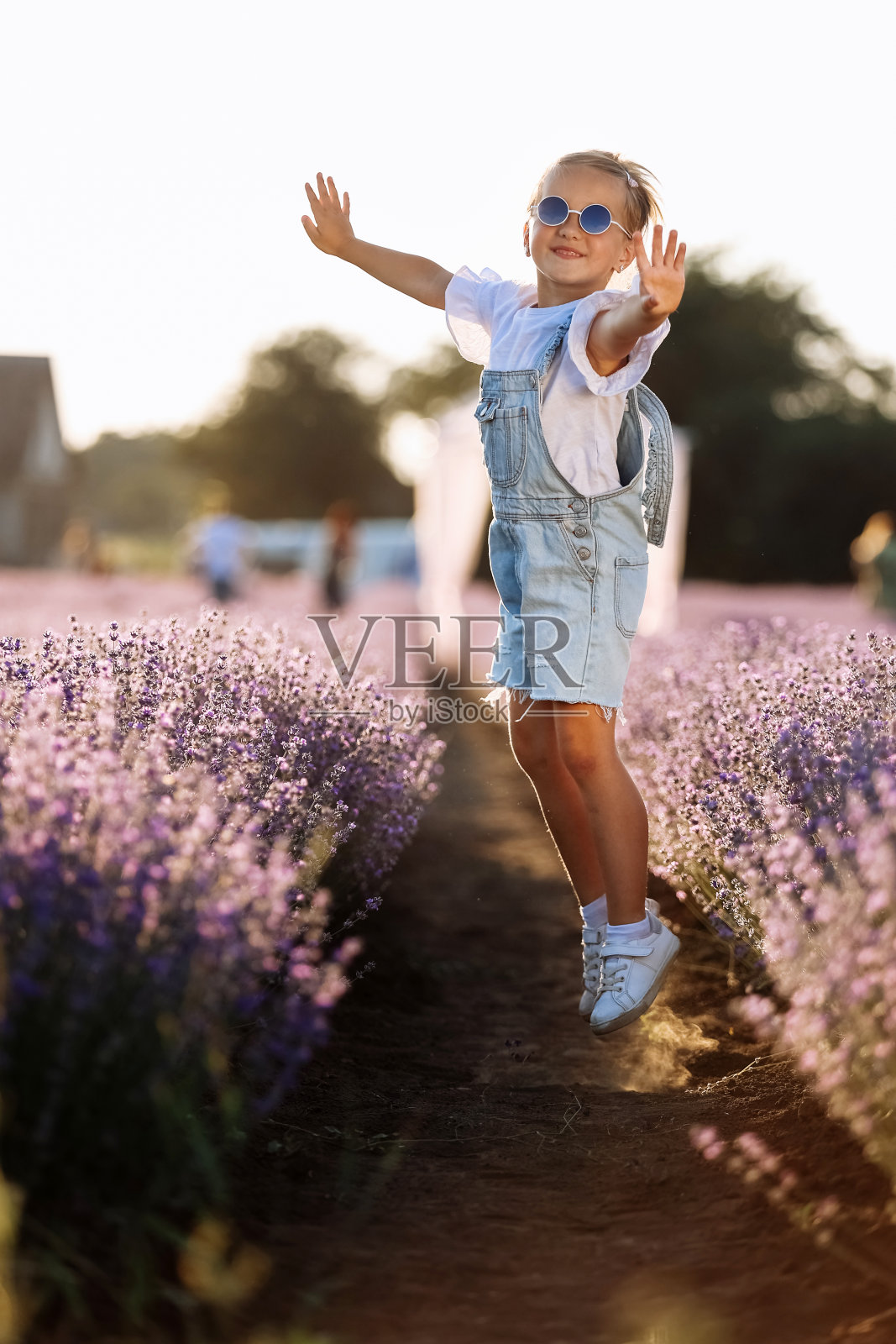 可爱的快乐的小女孩在夏天温暖的日子里在薰衣草地里玩耍和跳跃。戴着太阳镜，穿着牛仔裤连身裤，在大自然中微笑着的过度活跃的小孩。快乐童年概念照片摄影图片