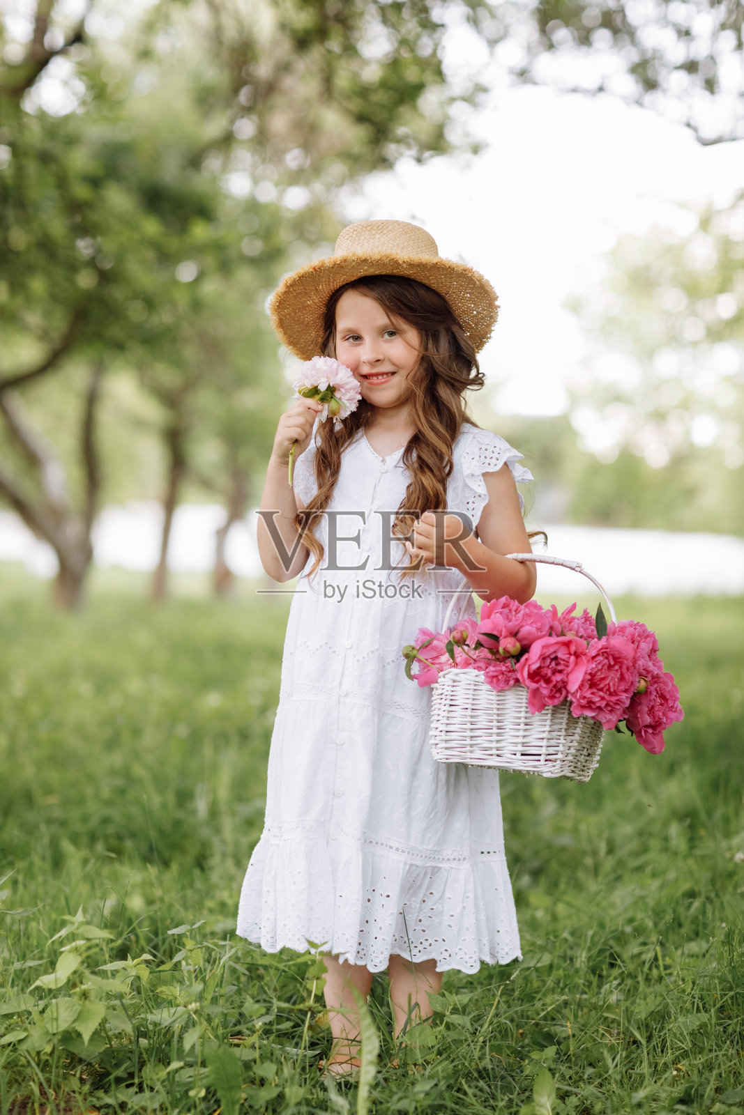 小女孩穿着白色蕾丝裙，头戴草帽，面带微笑，手拿柳条花篮，捧着粉红色的牡丹花，在绿色的公园里散步，享受夏日，温暖，鲜花，自由。照片摄影图片