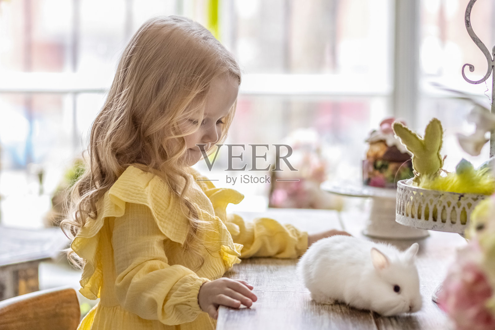 一个穿着黄色连衣裙的金发小女孩和兔子一起坐在复活节的餐桌旁。一个婴儿和一只兔子。复活节的概念。复活节在餐桌上。基督教的传统。照片摄影图片