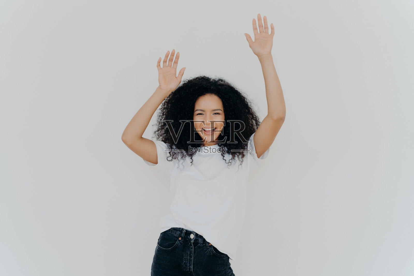 半长镜头，精力充沛、无忧无虑的卷发女人举手欢呼，穿着白色t恤和牛仔裤，有幸运的一天，完美的周末，白色背景下的模特。大家都举起手来照片摄影图片