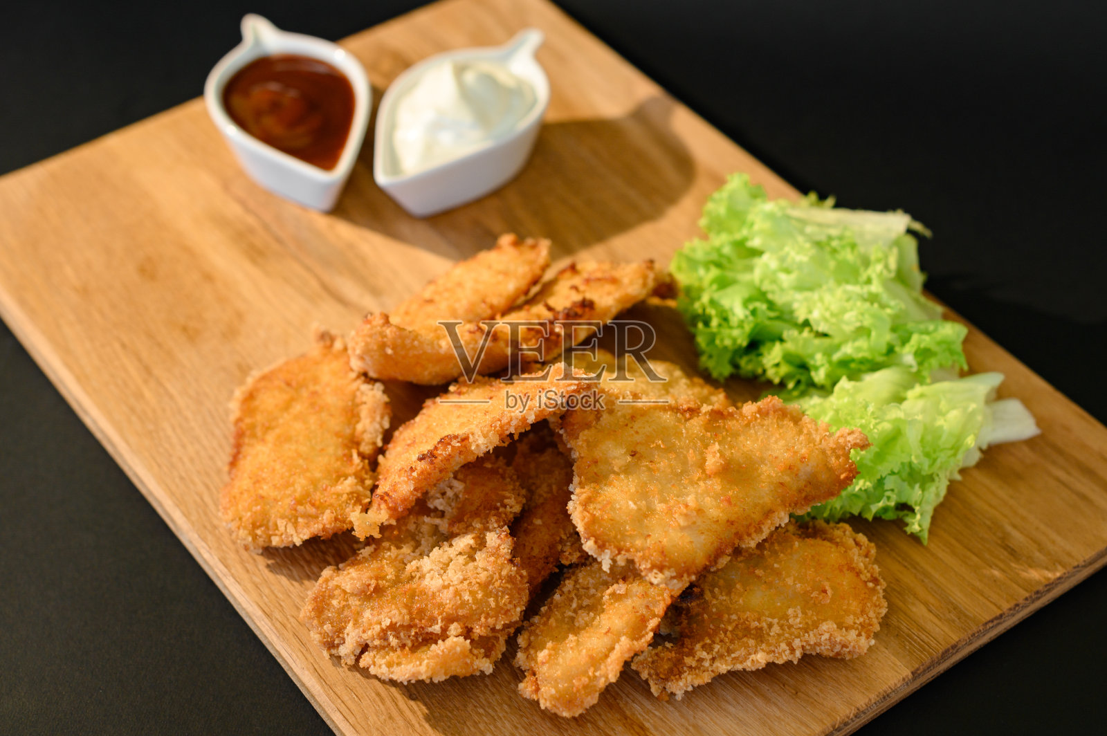 鸡块和酱汁，蛋黄酱和番茄酱，黑色背景的木板和沙拉叶。照片摄影图片