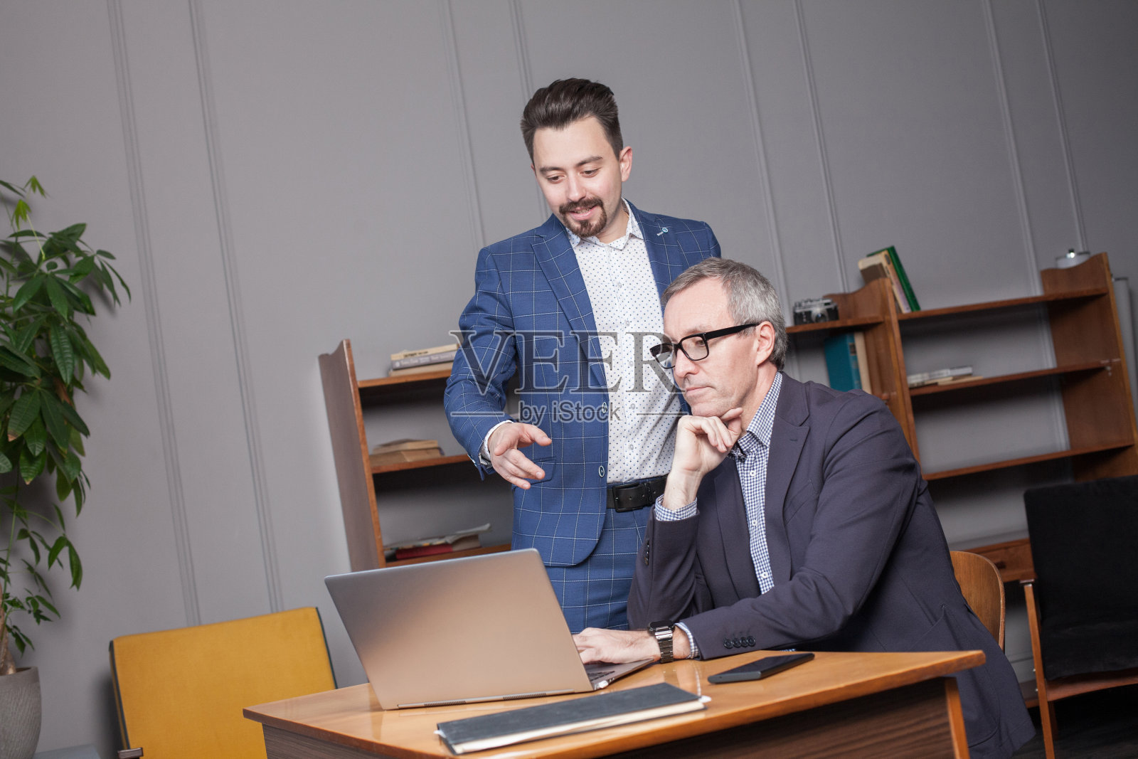 穿着蓝色西装，戴着眼镜，沉思的成熟商人在办公室里通过笔记本电脑与同事交谈照片摄影图片