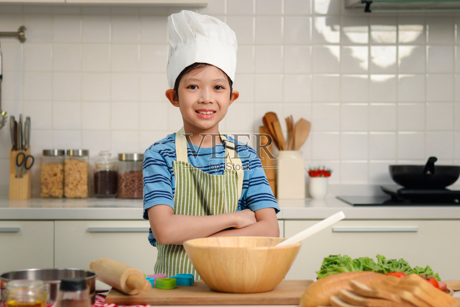 幸福微笑的亚洲男孩戴着围裙和厨师帽，双手站在厨房，面包师可爱的孩子厨师烹饪和准备烤面包，孩子自己做饭的肖像。照片摄影图片
