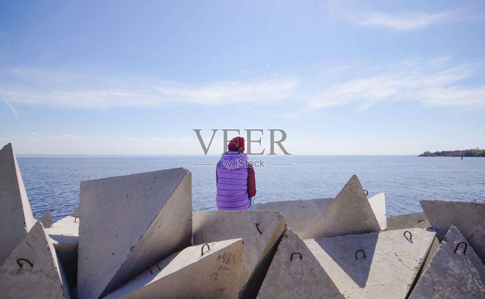 在一个阳光明媚的春日，一位戴着帽子的年轻女子坐在防波堤上欣赏大海。防波堤上穿着保暖衣服的女孩。照片摄影图片