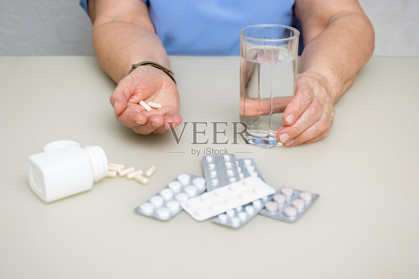 老妇人坐在桌旁，老皱的手里拿着白色的胶囊，维生素或治疗药片和水杯，保健和医药的概念照片摄影图片