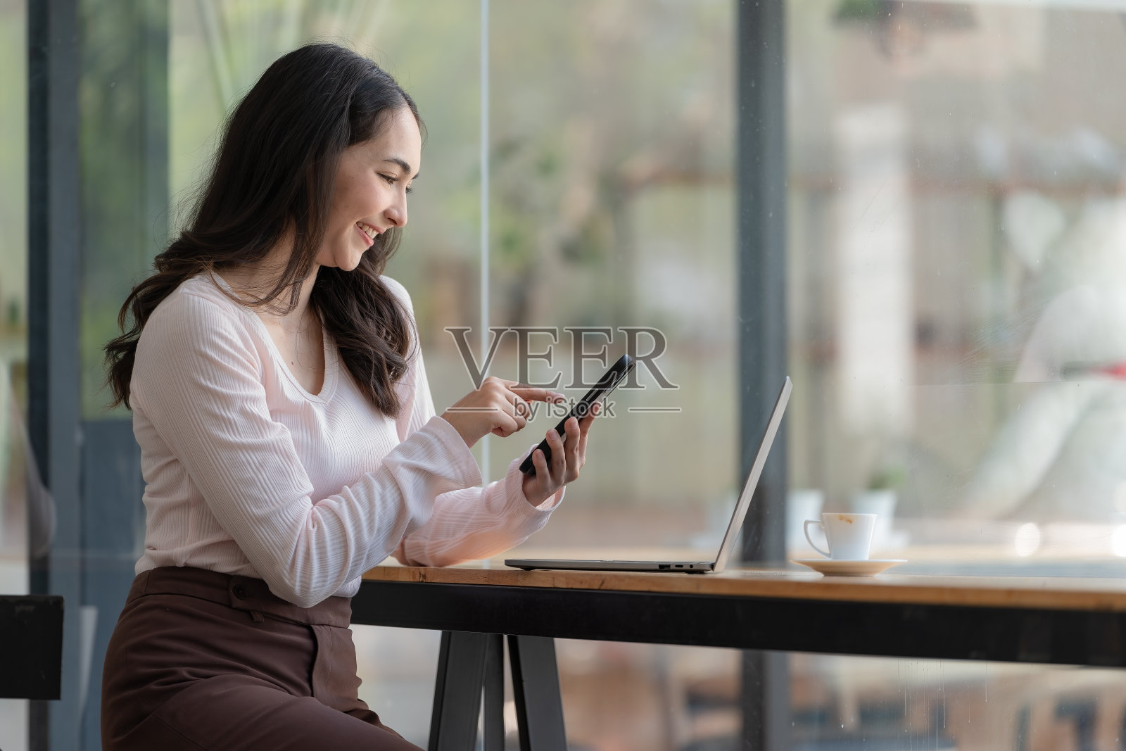 一个快乐的年轻亚洲女人坐在她工作的地方，在咖啡馆里讲着手机，面带微笑。照片摄影图片
