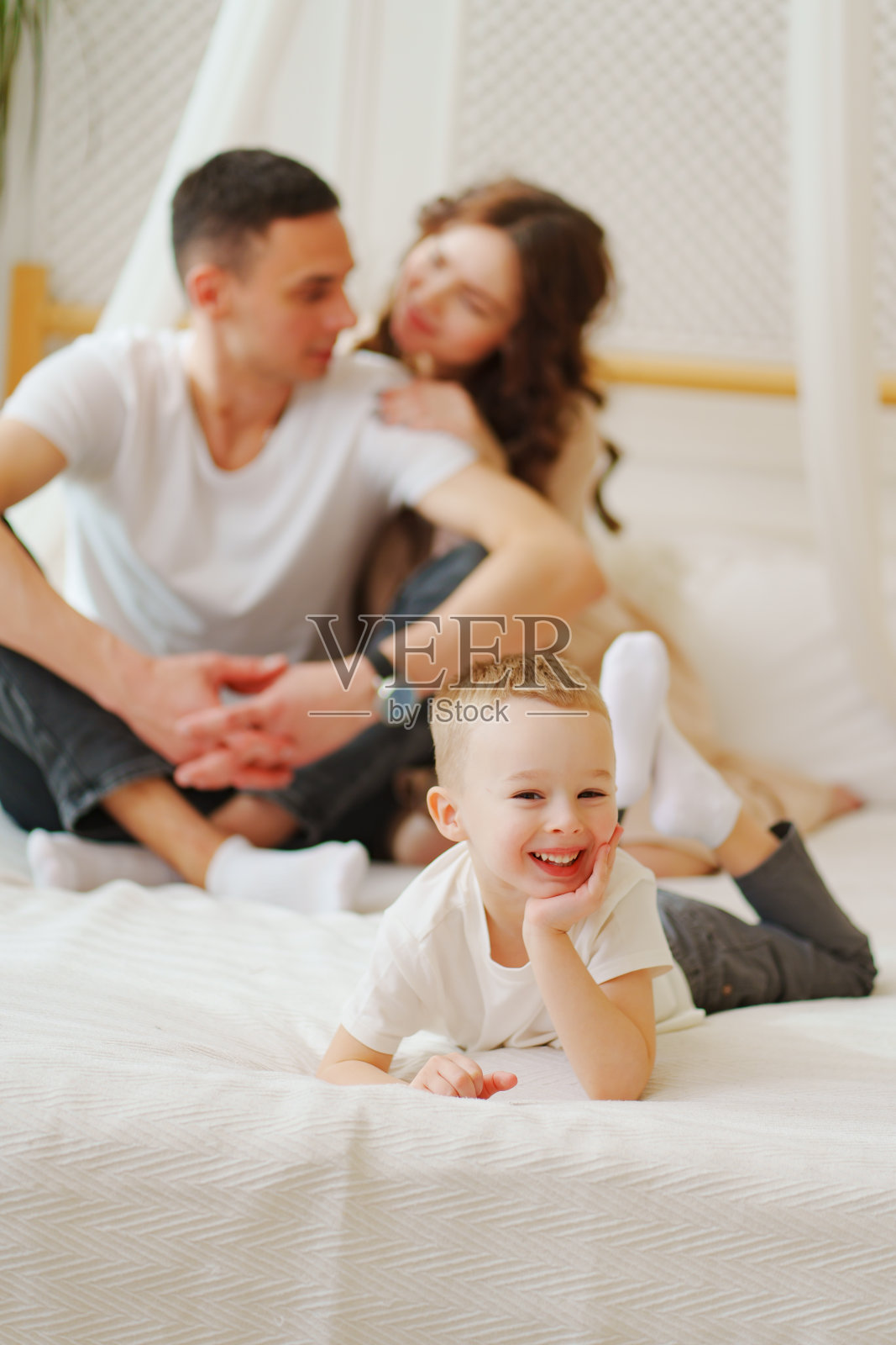 有趣的小儿子躺在床上，背景是慈爱的爸爸妈妈照片摄影图片