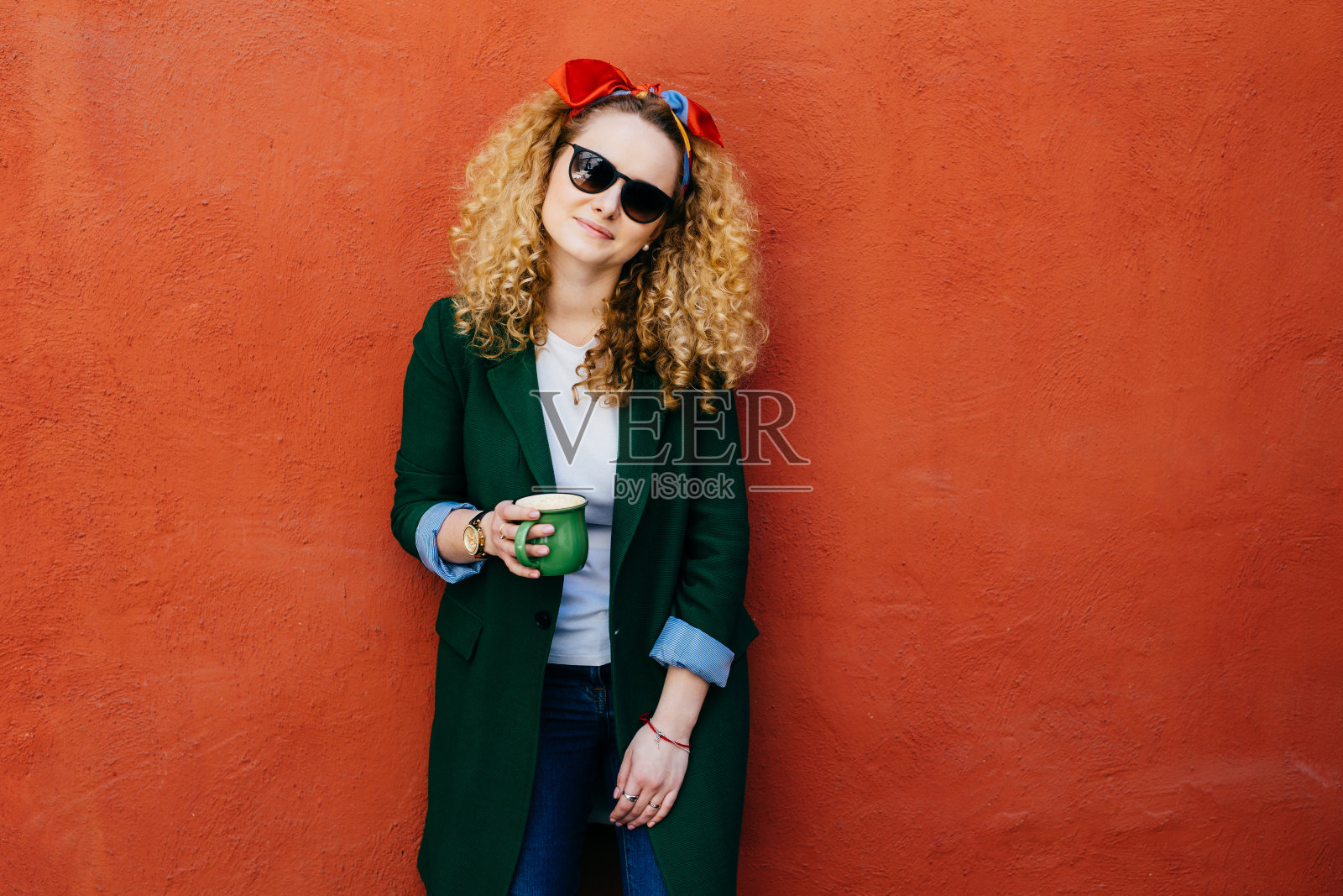 人与休闲的理念。漂亮女孩戴着发带、夹克、牛仔裤和太阳镜的横向肖像，拿着一杯热饮，喝茶或咖啡，在橙色的背景下摆姿势照片摄影图片