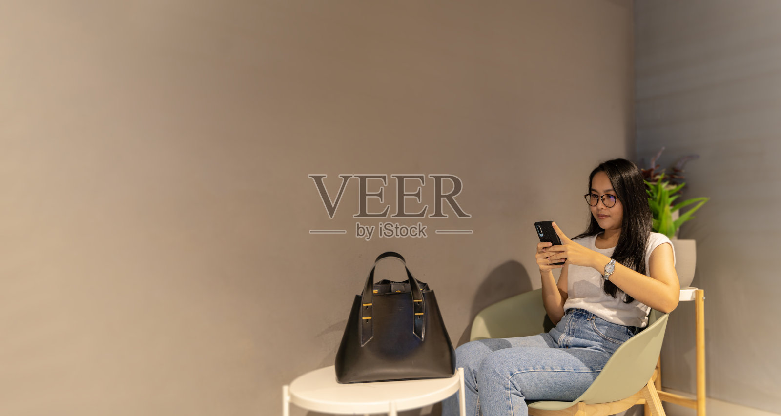 时尚的亚洲女性坐在咖啡馆里，喝咖啡，用智能手机，在户外看视频。背景墙是空的，可以放文字照片摄影图片