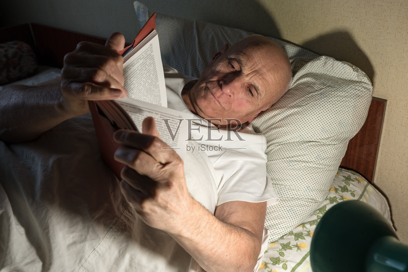 老人独自一人躺在床上，在电灯下看书照片摄影图片
