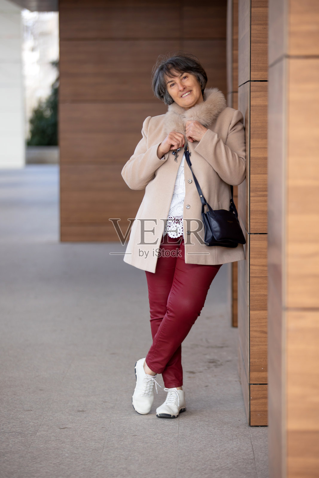 穿着漂亮外套的老妇人，站在墙边，看着相机，女性肖像照片摄影图片