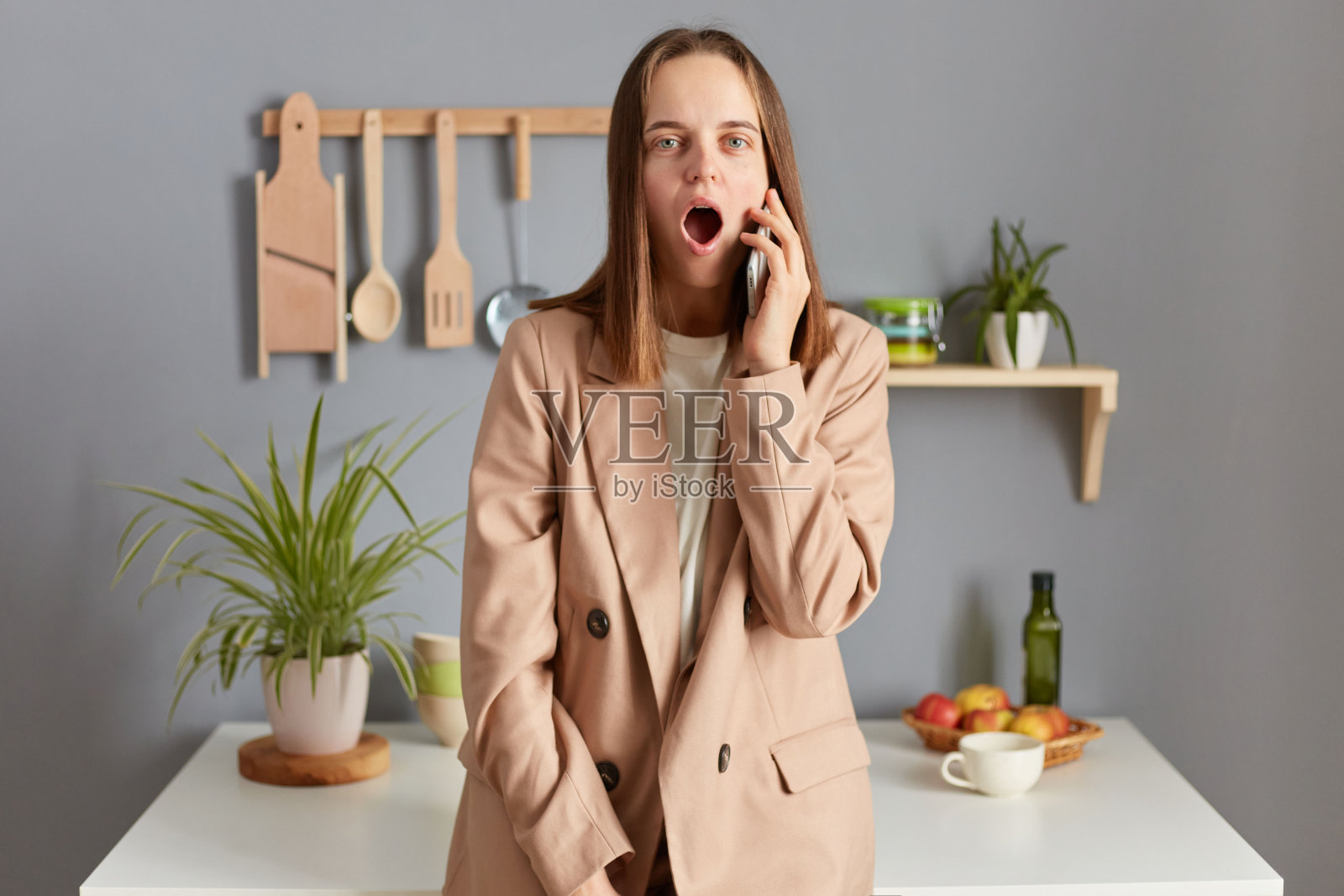 震惊的白人女性穿着米色的正装站在厨房的桌子旁边，用手机聊天，听到可怕的消息，嘴巴张得大大的。照片摄影图片