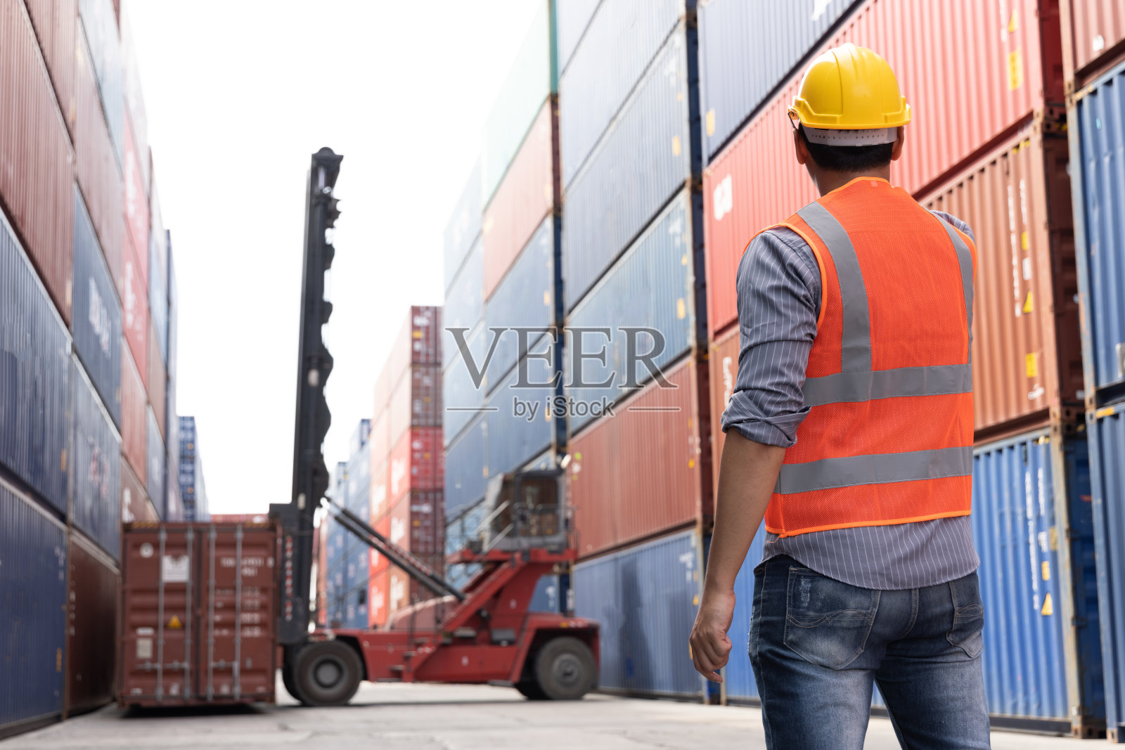 在集装箱仓库工作的一名工人站在货场看着叉车搬运红色集装箱照片摄影图片