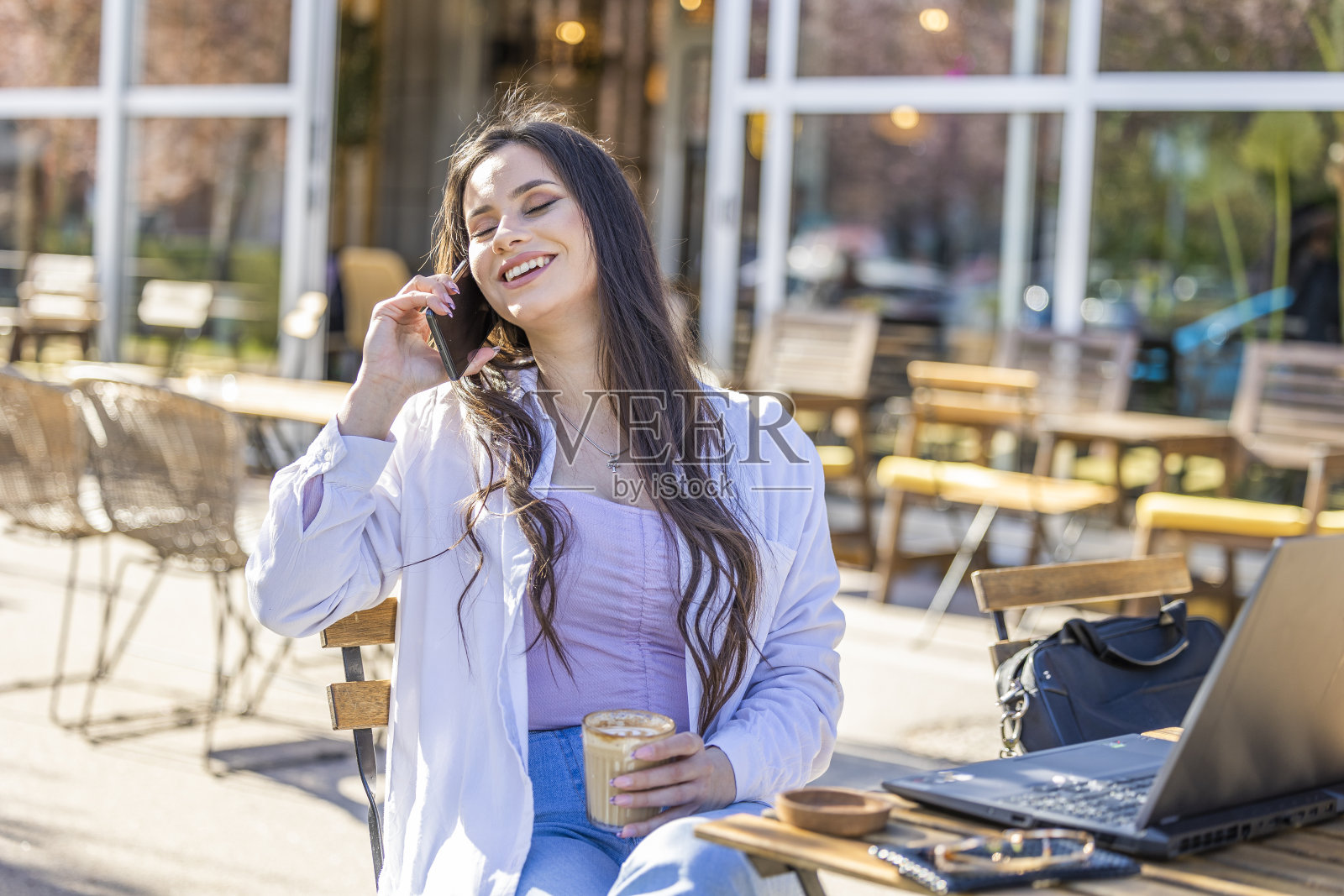 Beautiful girl giggles while talking on the phone and holding a coffee cup in her hand at the sidewalk café照片摄影图片