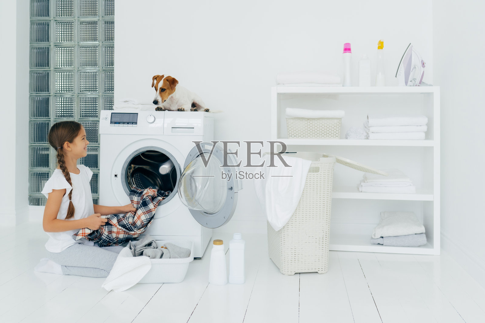 积极的女孩清空洗衣机，拿着干净的格子衬衫，微笑着看着最喜欢的宠物，它帮着洗衣服，在白色的地板上摆姿势，盆里装满了衣服和清洁剂。照片摄影图片
