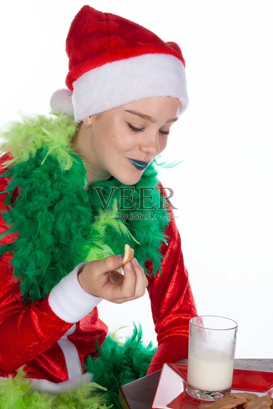 小女孩的特写肖像戴着红色圣诞老人帽像圣诞怪杰厚颜欢笑吃饼干看着牛奶，孤立的白色背景。有趣的人类情感照片摄影图片