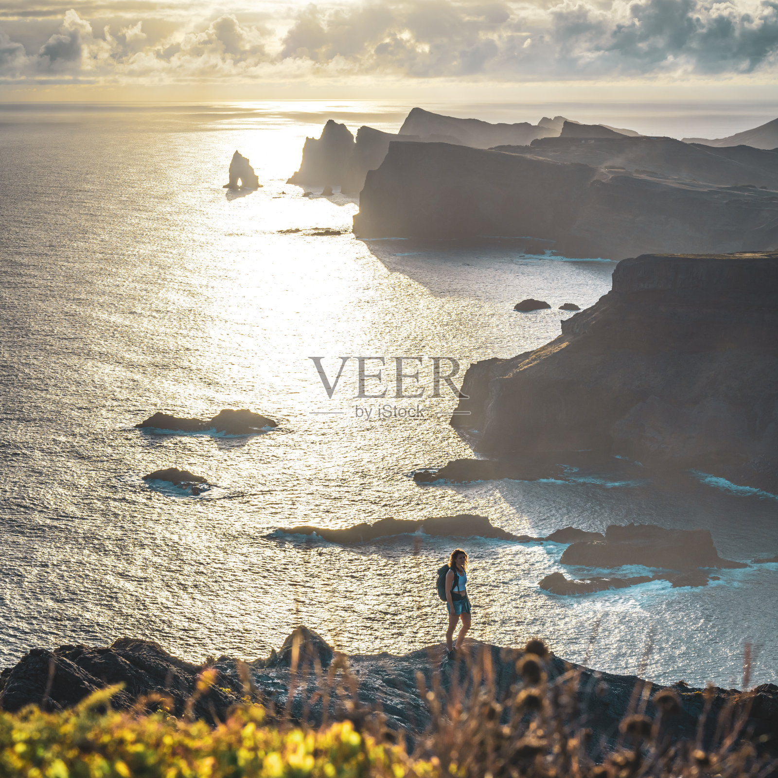 背包客妇女喜欢徒步旅行在一个陡峭的悬崖上的海景和沿着崎岖的山麓马德拉海岸在日出。Ponta do Bode，马德拉岛，葡萄牙，欧洲。照片摄影图片