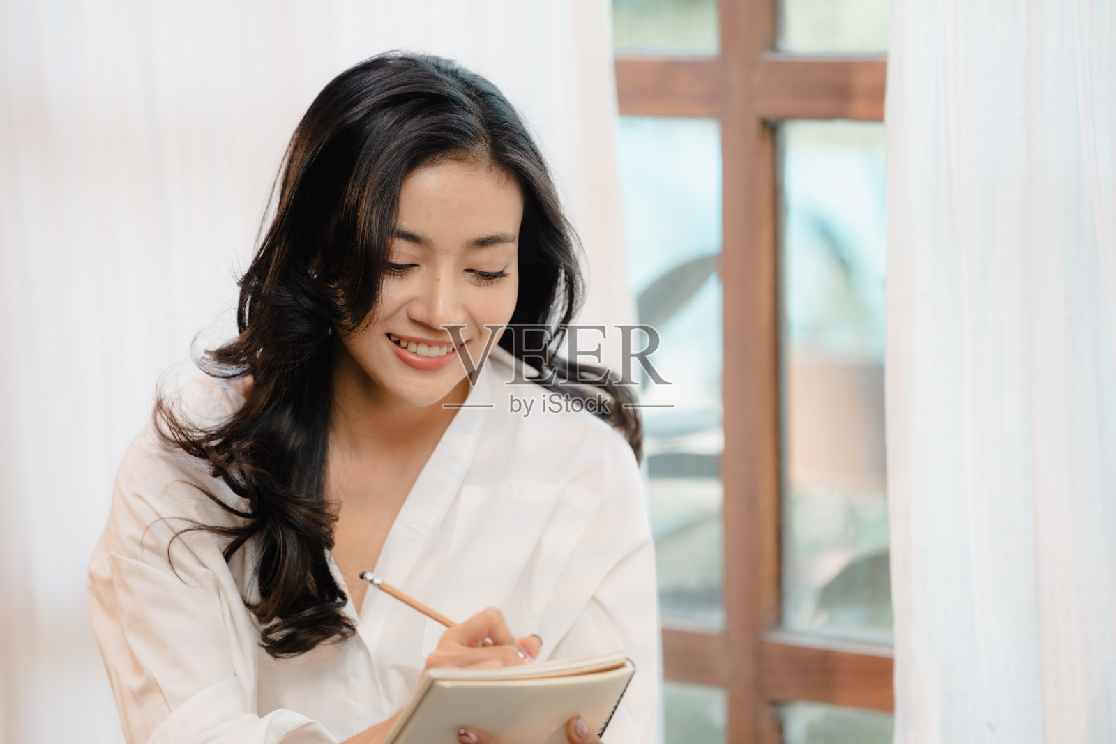 漂亮的年轻亚洲女人穿着白色连衣裙躺在沙发床上写日记。微笑的黑发女人手里拿着笔记本和笔。现代明亮舒适的家庭内部背景。照片摄影图片