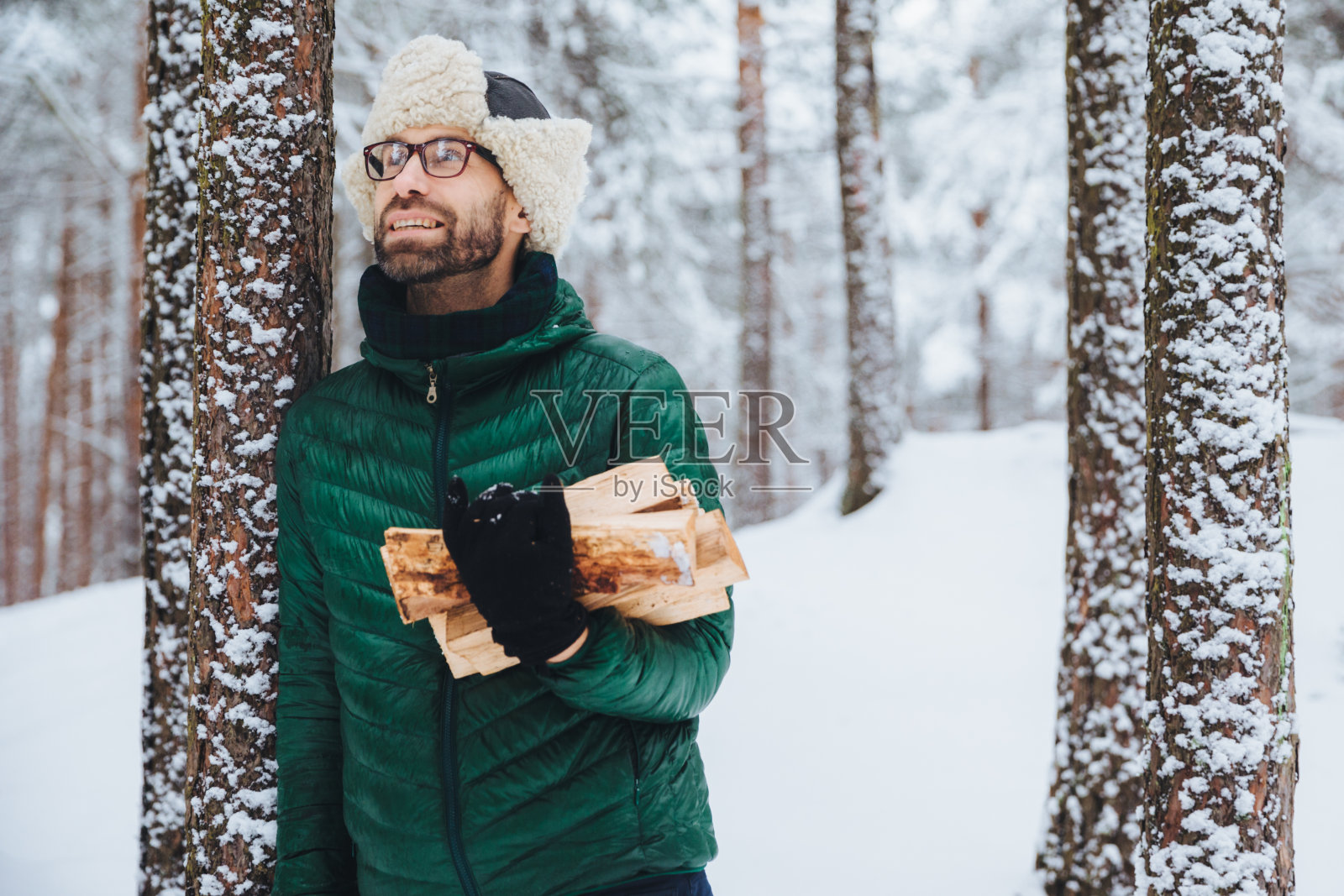 户外微笑的男性肖像站在被雪覆盖的树旁，拿着柴火，高兴地抬头看着树上的松鼠，站在冬天的森林里。人与娱乐理念照片摄影图片