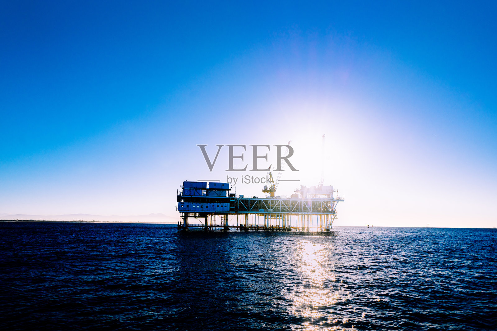 当太阳从浩瀚的太平洋升起时，一座海上石油钻井平台矗立在地平线上。钻井平台高耸的轮廓在温暖的晨光下熠熠生辉。照片摄影图片