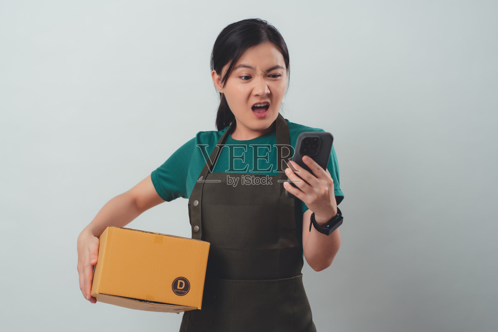 身穿围裙，拿着纸箱，用智能手机与客户发生冲突的亚洲女性。照片摄影图片