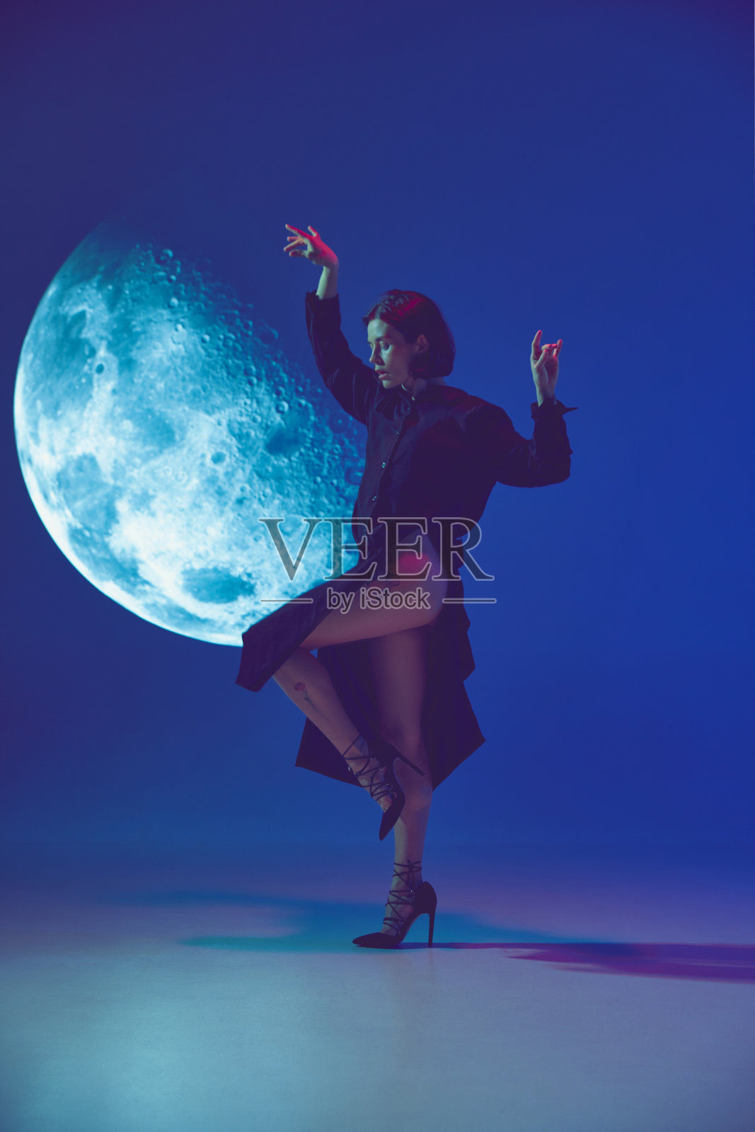 一个梦中的女人穿着优雅的连衣裙，在霓虹灯的蓝色工作室背景下，在投影附近摆姿势。满月照片摄影图片