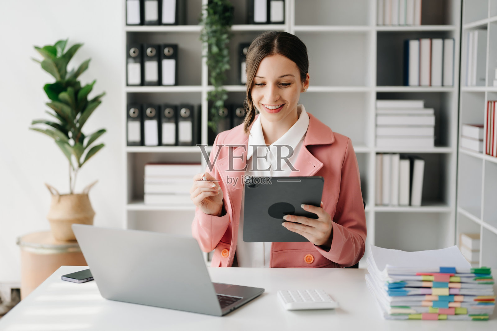 自信的商业专家迷人的微笑的年轻女子在办公室的桌子上打字笔记本电脑和拿着数字平板电脑。照片摄影图片