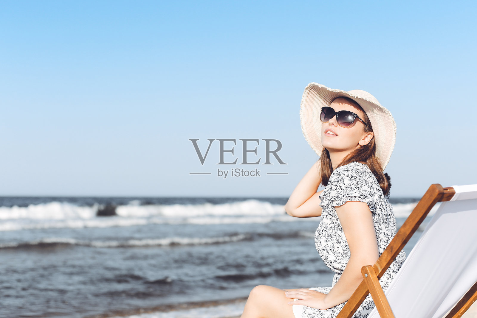 Happy brunette woman wearing sunglasses while relaxing on a wooden deck chair at the ocean beach照片摄影图片