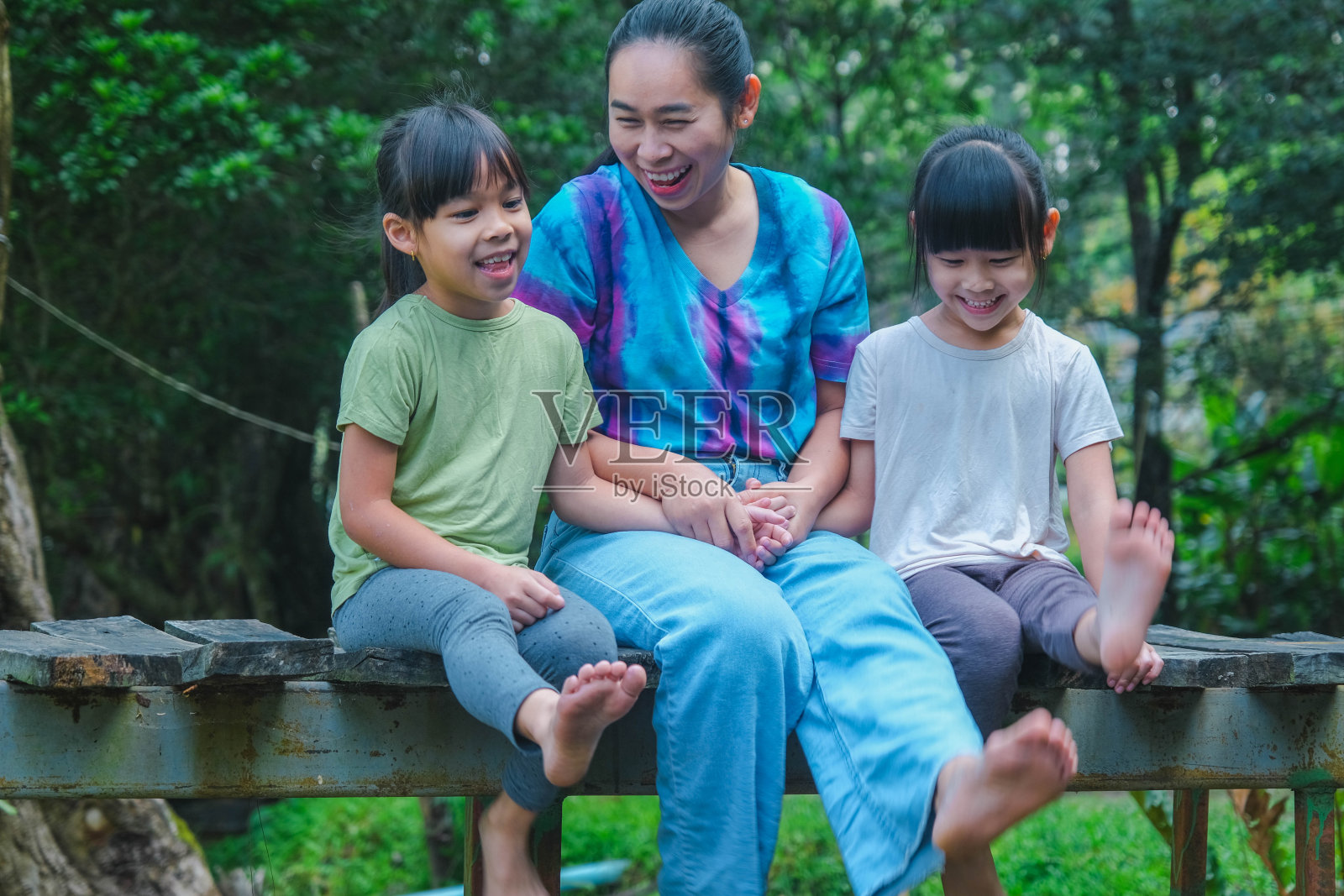 夏天，快乐的年轻母亲带着几个小女孩坐在河边的木桥上。母亲和两个女儿在户外玩得很开心。祝充满爱的家庭和母亲节快乐。照片摄影图片