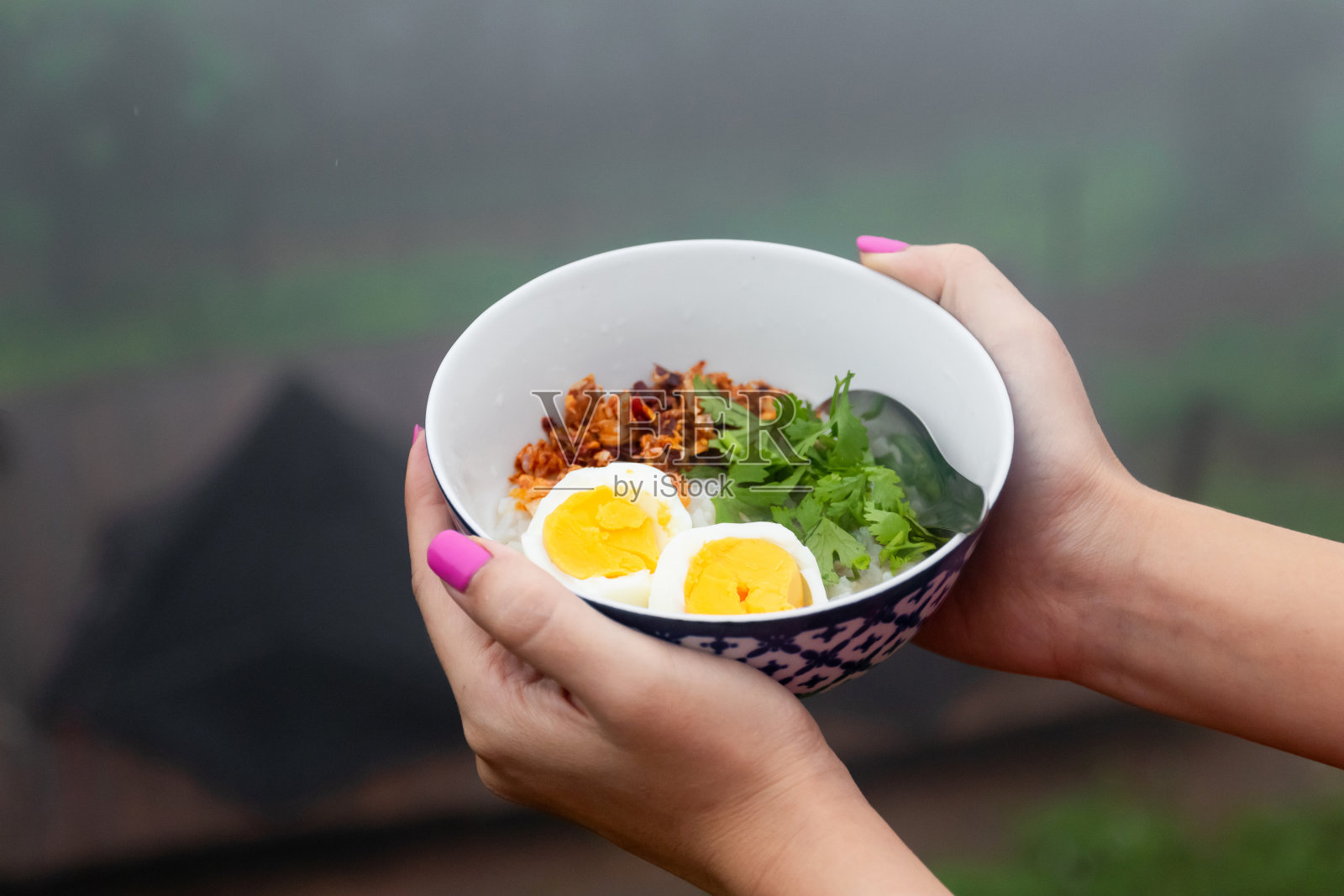 女人手里拿着一碗米粥和煮鸡蛋，炒大蒜、辣椒和香草。雨季有晨雾的早餐。照片摄影图片