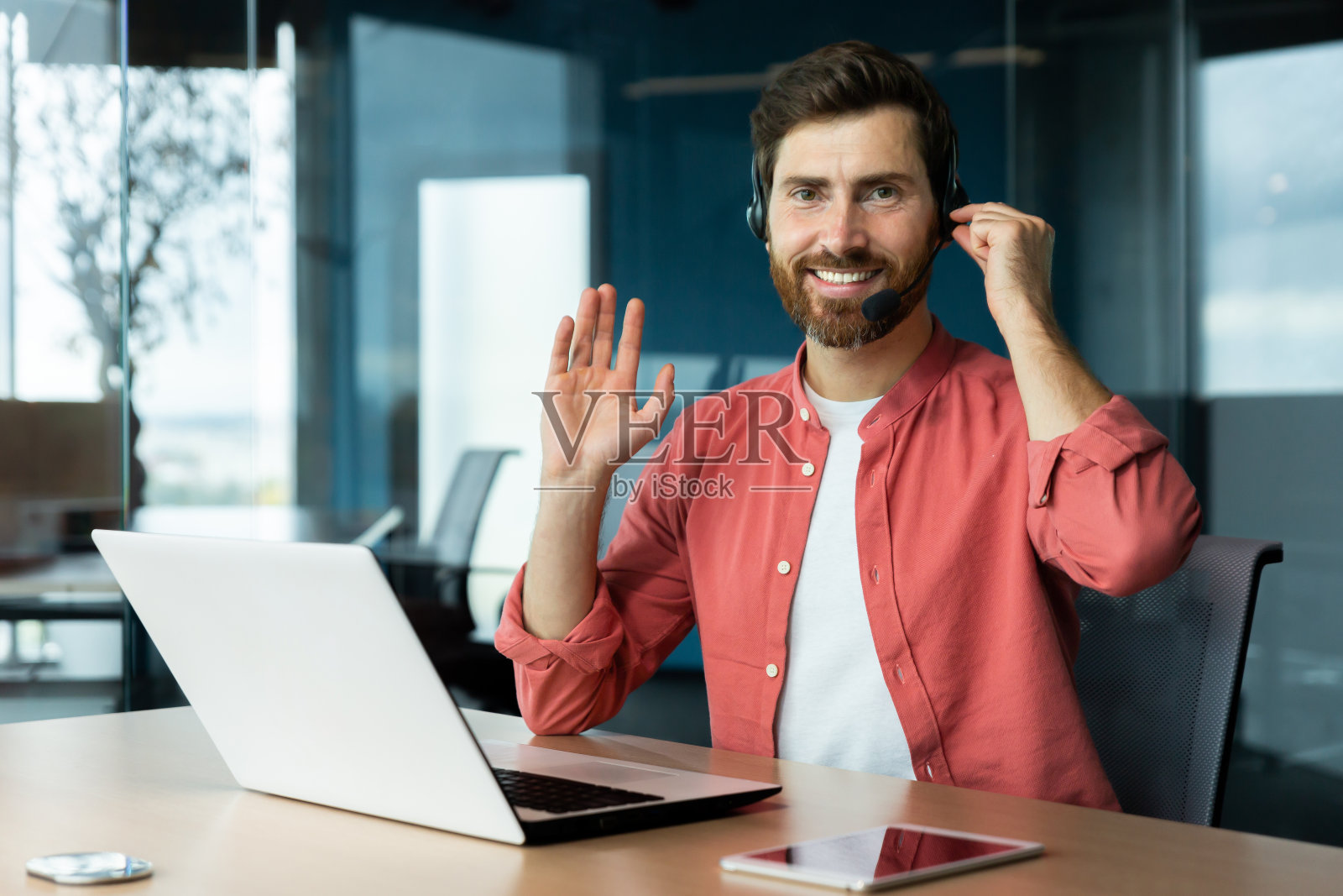 一个戴着心理学家，心理治疗师耳机的年轻人，坐在办公室的桌子旁，拿着笔记本电脑，在网上咨询，进行讲座，看着并问候镜头，微笑着照片摄影图片
