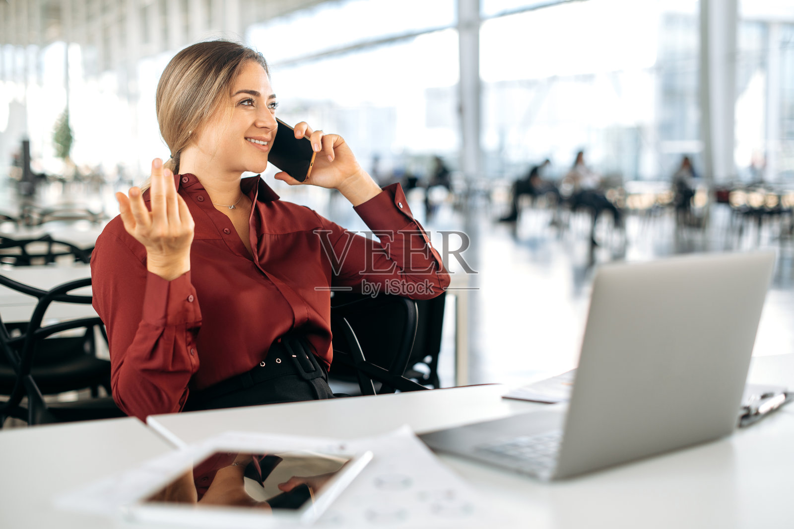 积极美丽优雅的金发白种女人穿着正式的衬衫，办公室职员，经理，坐在办公室的办公桌前，与同事或客户用智能手机聊天，看向别处，微笑照片摄影图片