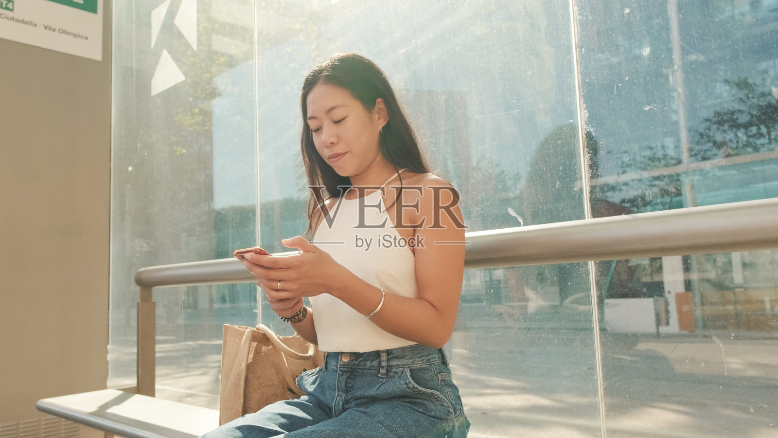 漂亮的黑发女孩，穿着白色上衣和牛仔裤，坐在公共交通站，手里拿着智能手机，环顾四周照片摄影图片