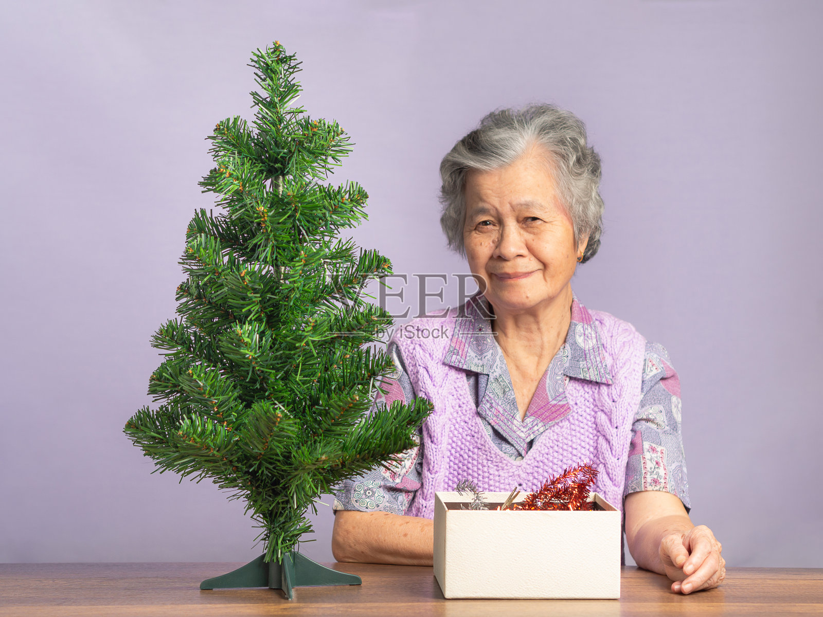 一位留着灰白短发的快乐老妇人坐在灰色的背景上准备装饰圣诞树。照片摄影图片