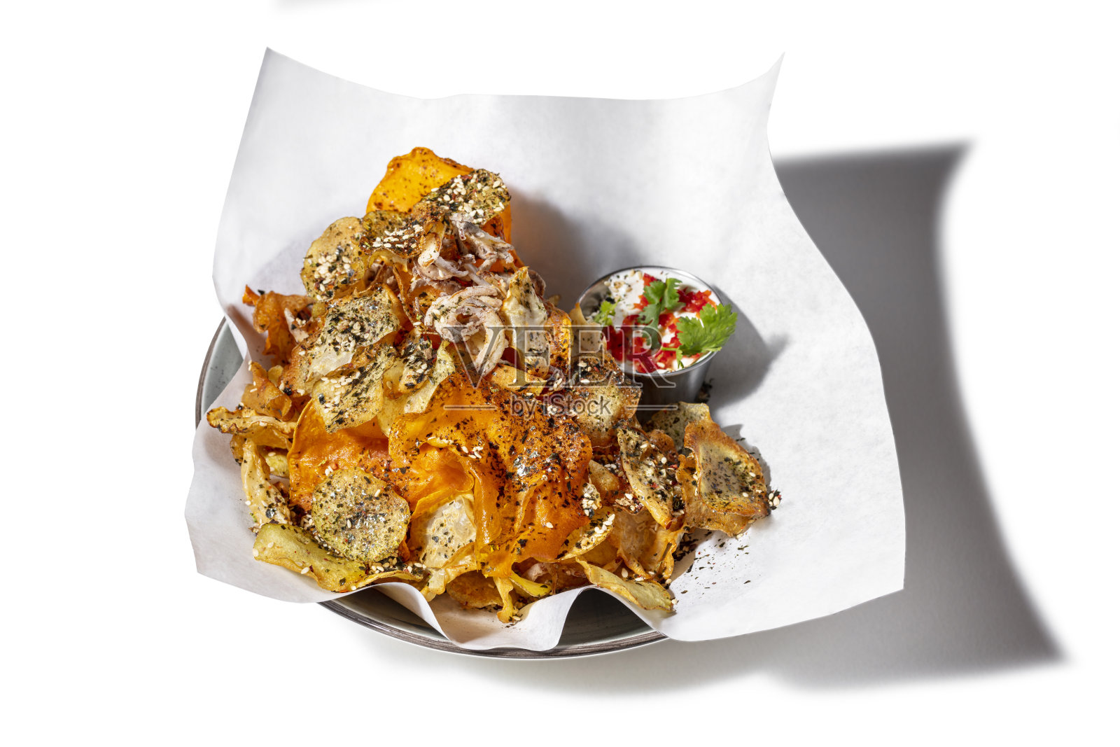 土豆南瓜片配香草和芝麻。食物放在一张白色的羊皮纸上，旁边是一个金属卤汁船，上面放着大蒜酱、切碎的辣椒和香菜叶。羊皮纸放在一个陶瓷盘子上。白色背景。照片摄影图片