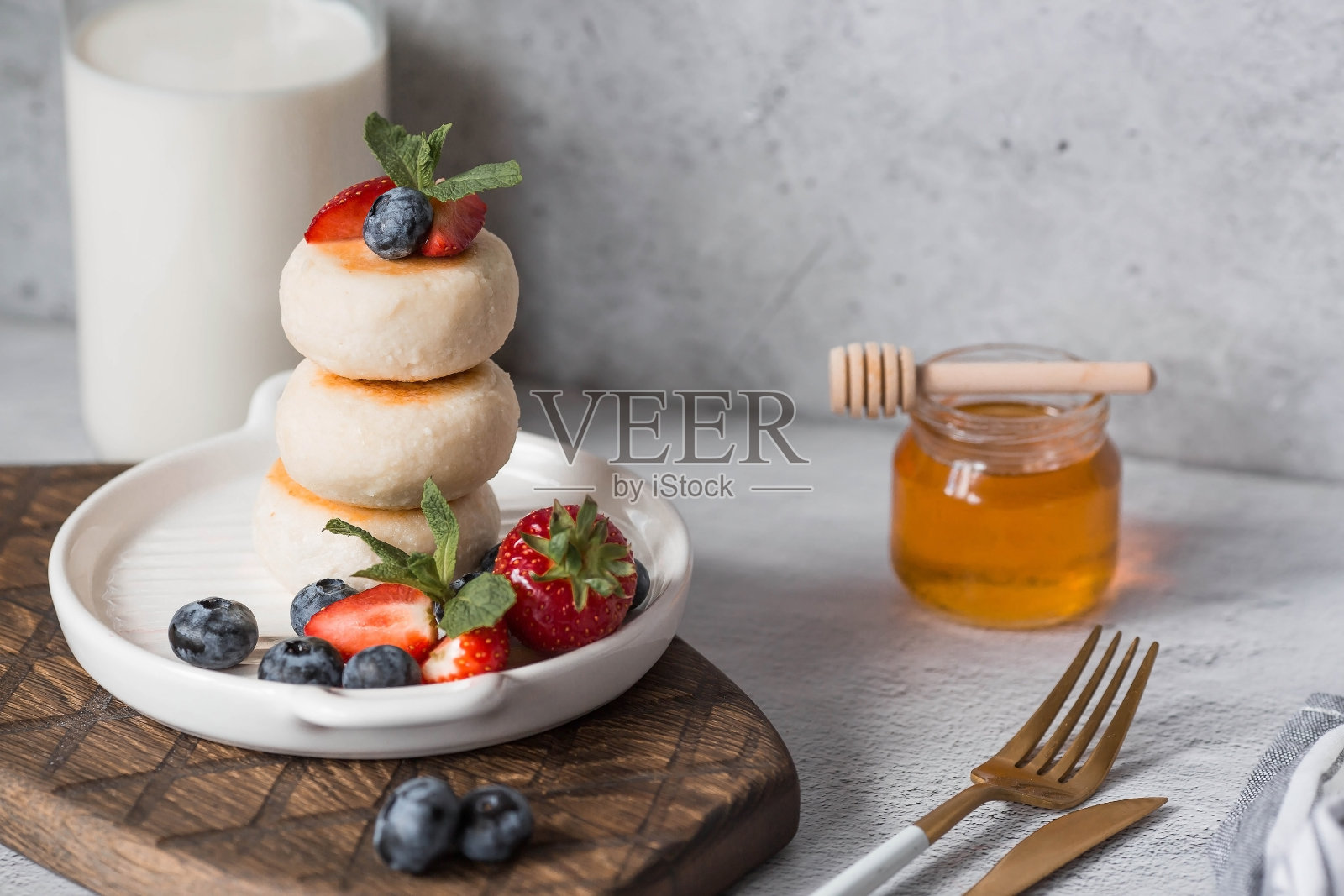 灰色混凝土桌子上时髦的木板上放着松软干酪煎饼、俄罗斯奶酪蛋糕和新鲜的蓝莓、草莓、蜂蜜和薄荷叶。自然产品。健康美味的早餐。照片摄影图片