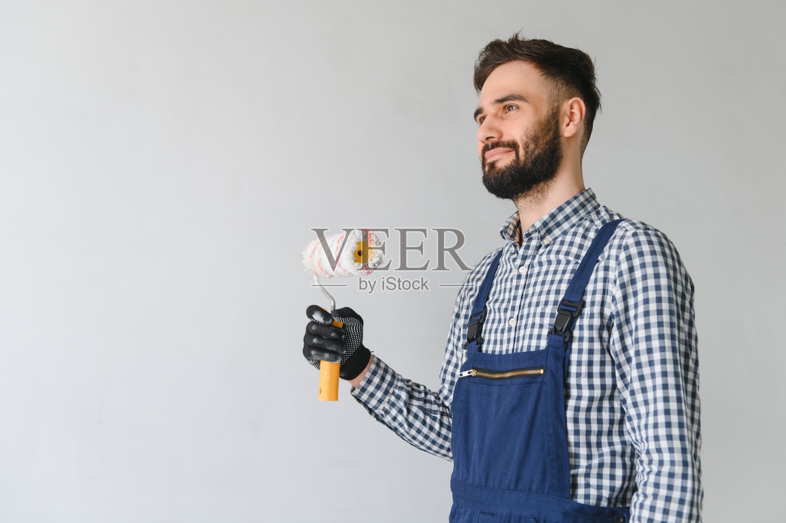 穿着蓝色制服的年轻专业工人微笑着站在新公寓的油漆滚筒上，在灰色的墙壁背景上进行修复，复制空间。照片摄影图片