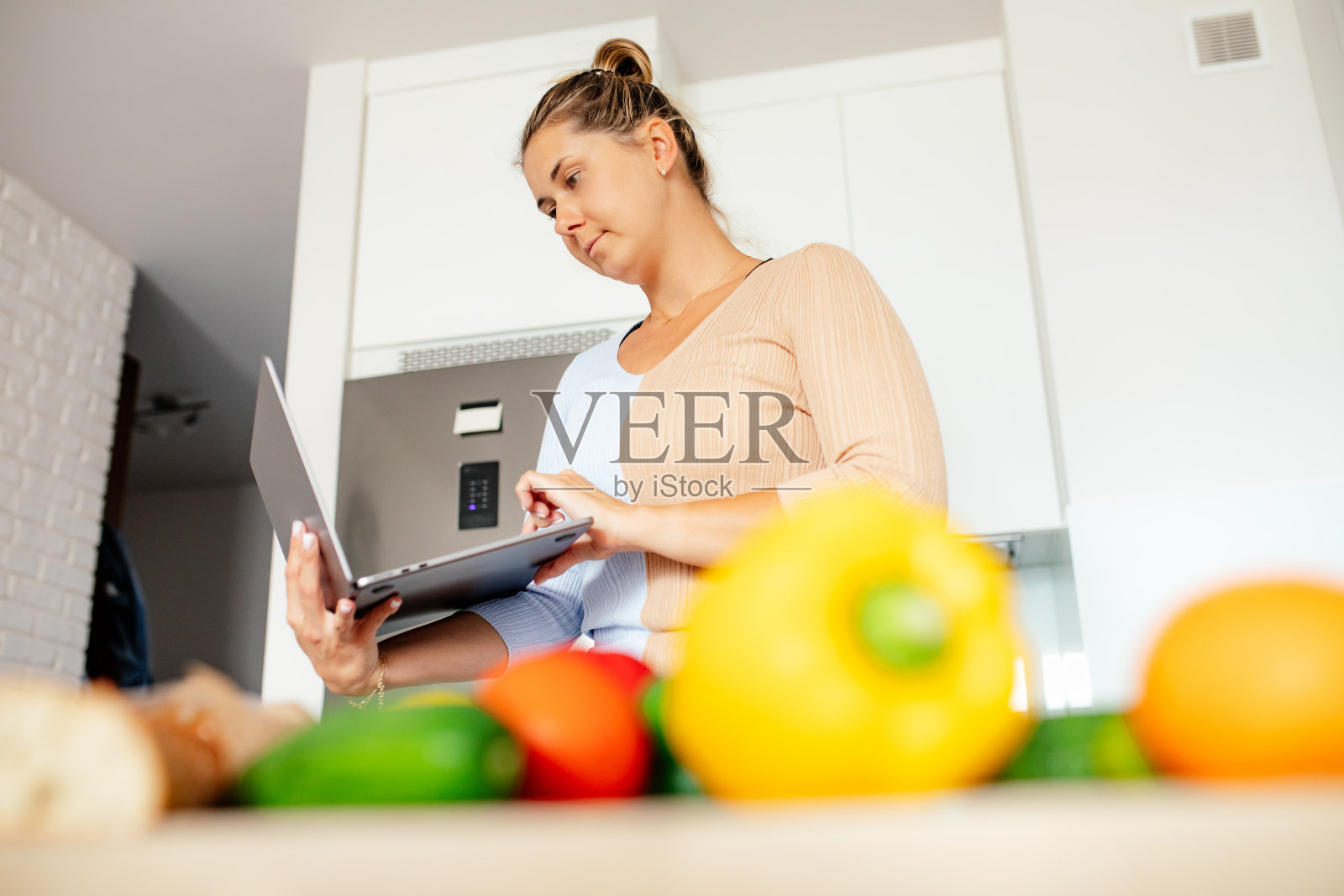 前景是未对焦的新鲜蔬菜，后面是一个拿着笔记本电脑寻找食谱的年轻女子。照片摄影图片