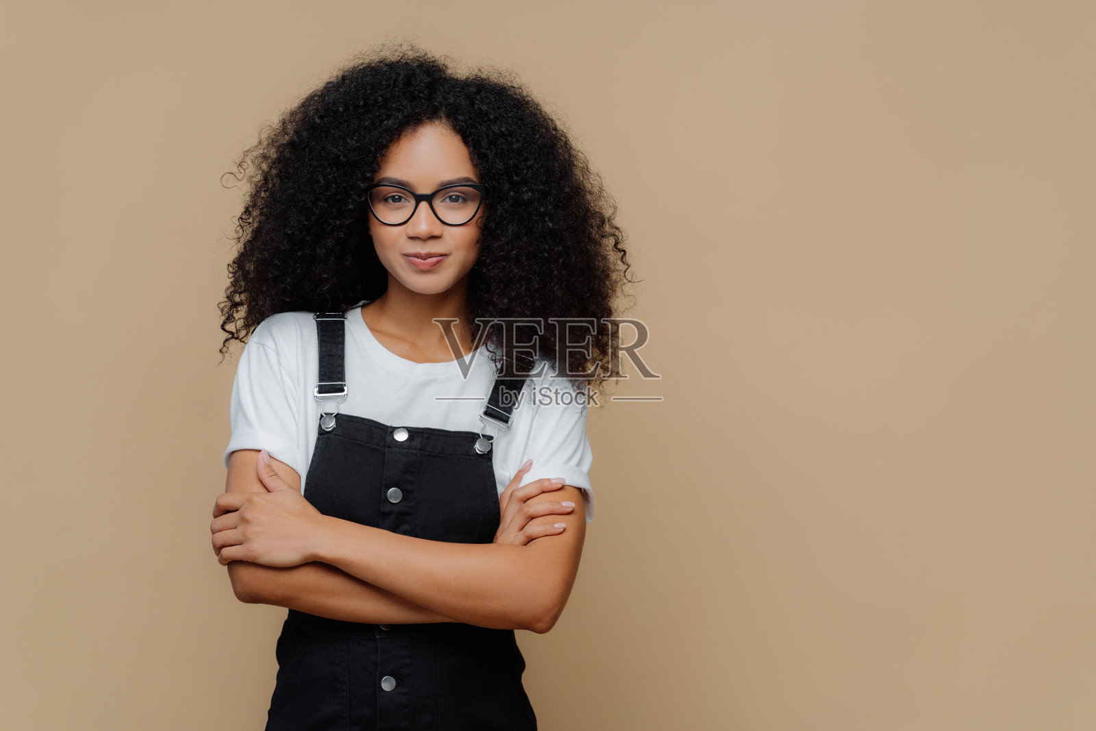 美丽的黑皮肤十几岁的女孩摆姿势，双臂交叉，戴眼镜，白色t恤和黑色沙拉芬，站在棕色的背景，复制空间为您的宣传内容照片摄影图片