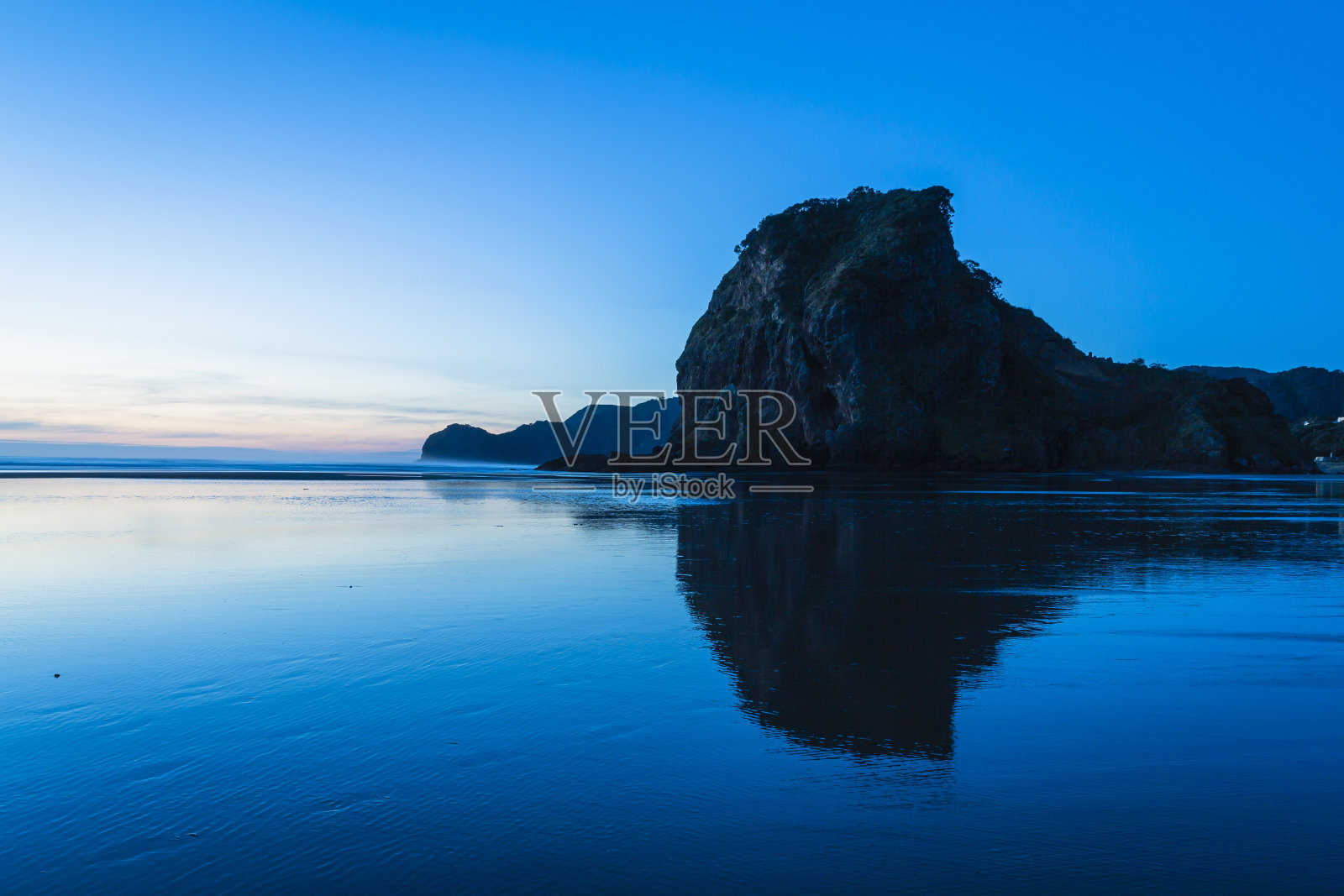 黄昏时分，在新西兰奥克兰附近的皮哈海滩，狮子岩映照在水面上照片摄影图片