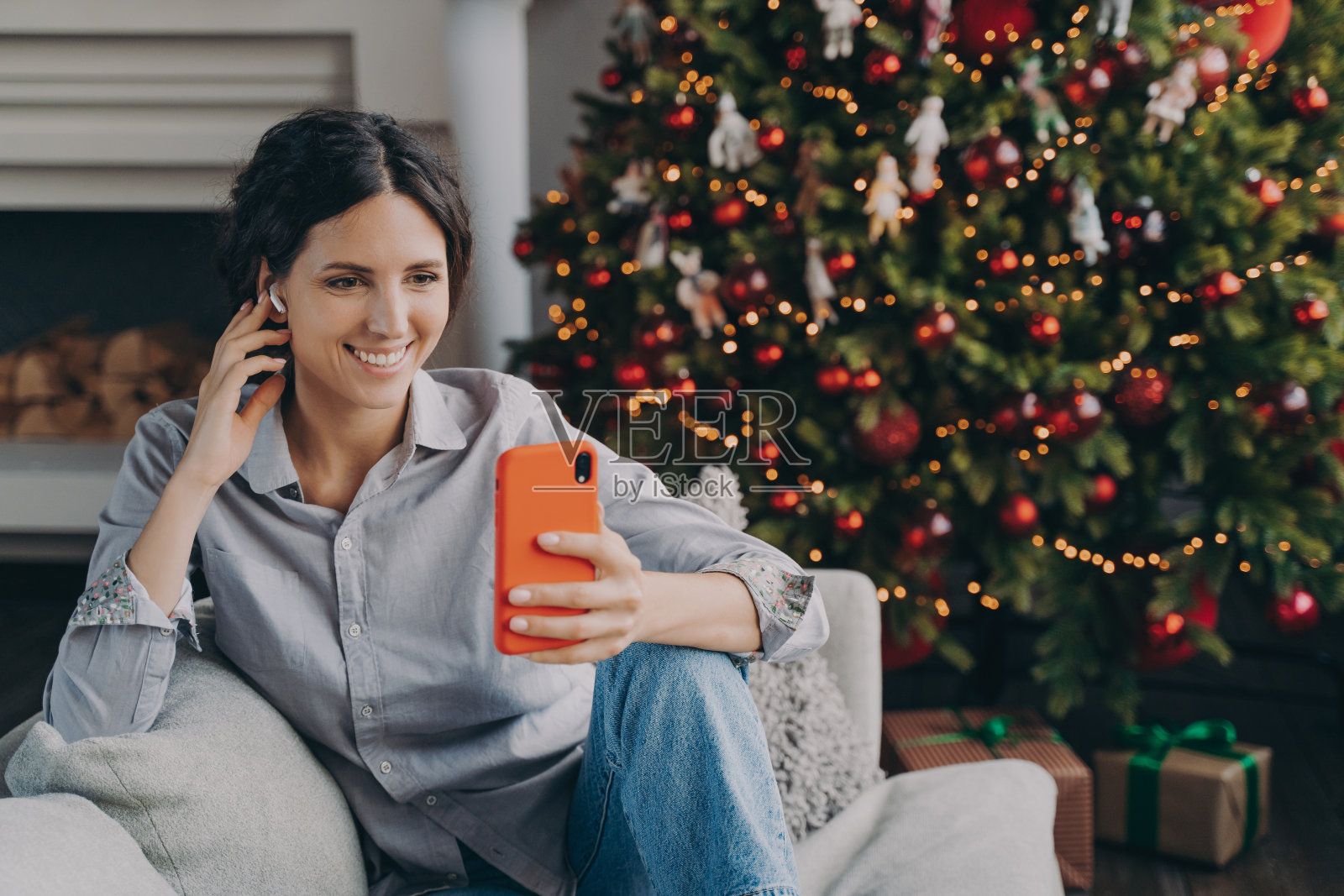 快乐的西班牙妇女戴着无线耳机，拿着电话，坐在家里的圣诞树旁照片摄影图片