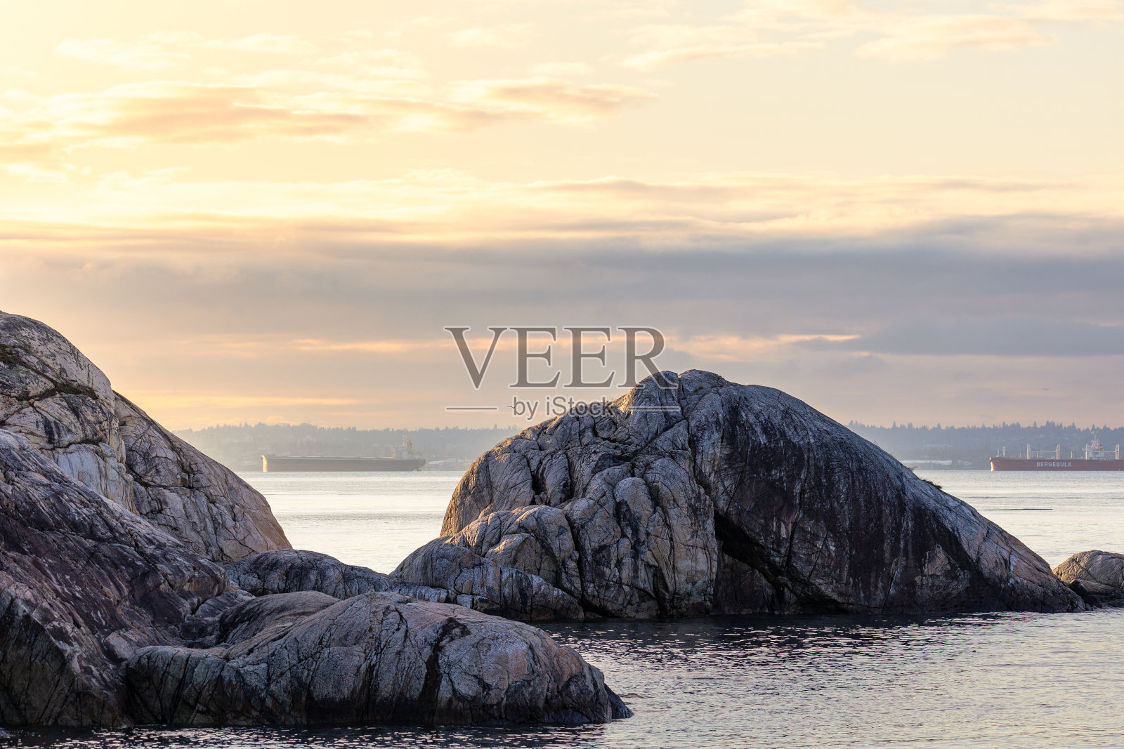 清晨朦胧的金色阳光洒在岩石嶙峋的海岸上，水面上有货船照片摄影图片