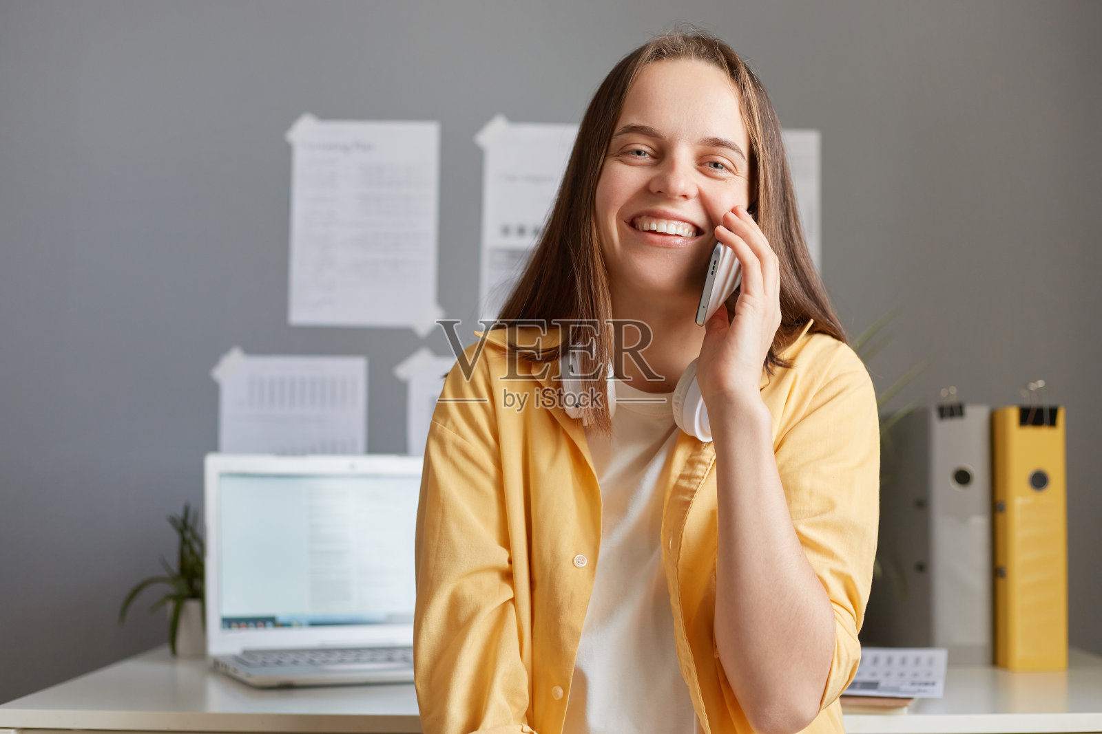 谈生意很愉快。穿着黄色夹克的年轻漂亮的时髦女人坐在工作的地方讲手机，微笑着看着镜头。照片摄影图片